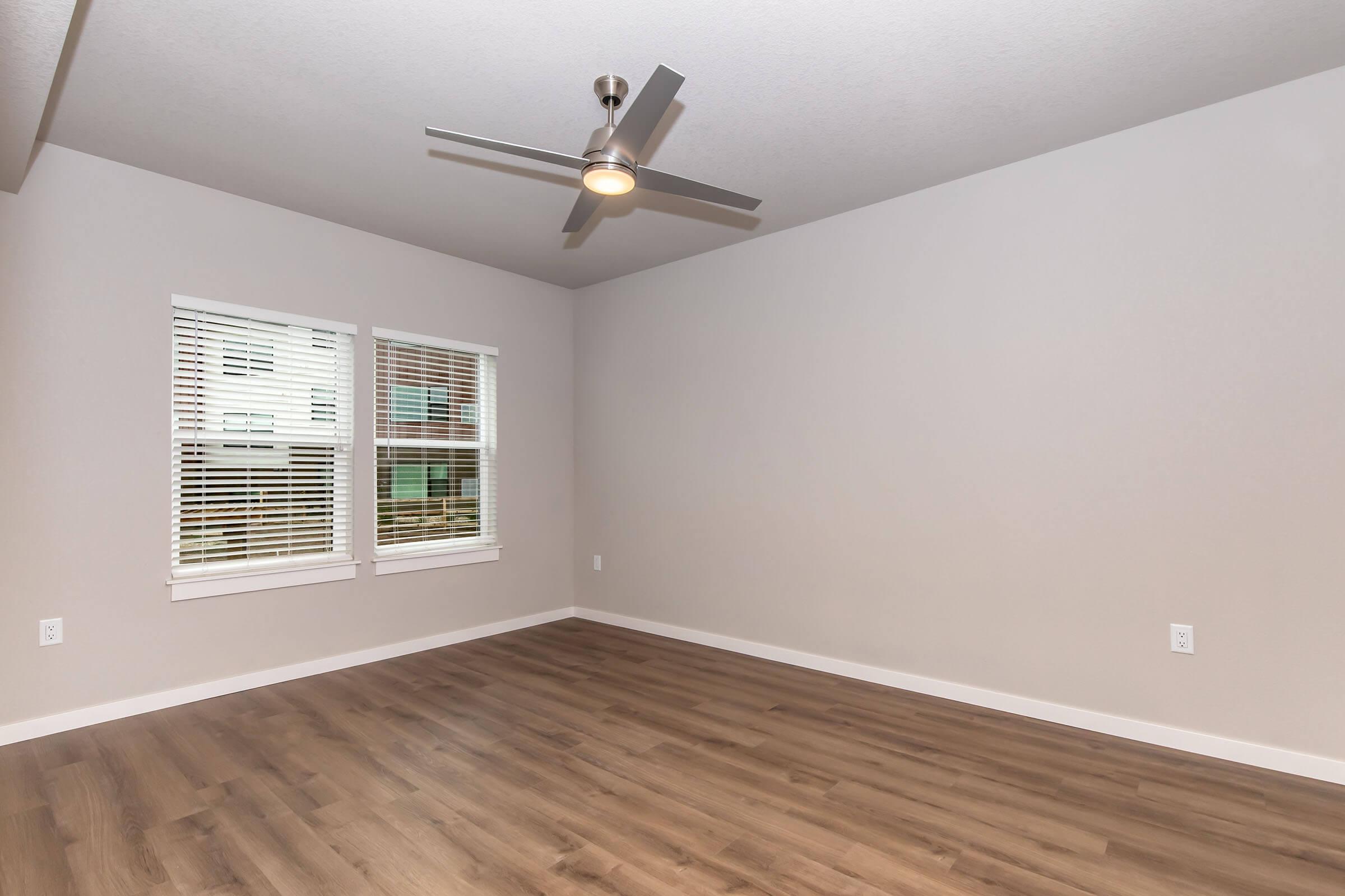 A spacious empty room with light gray walls and wooden flooring. There are two large windows with white blinds allowing natural light in. A modern ceiling fan is installed in the center. The decor is minimal, showcasing an open and airy atmosphere.