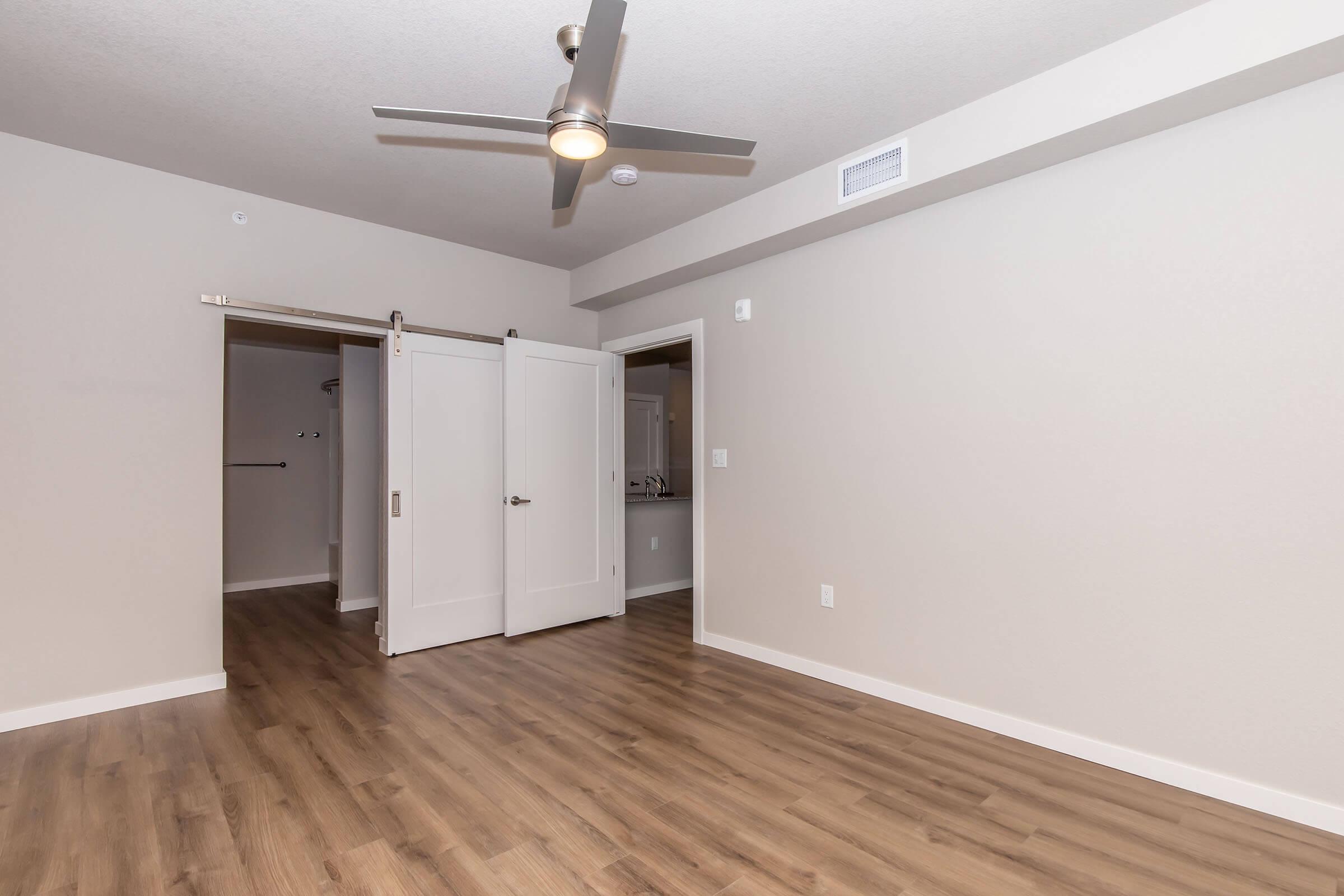 A spacious room with light-colored walls and wooden flooring. There are sliding double doors leading to another room, and a ceiling fan is visible. The room appears bright and empty, providing an open space with a modern aesthetic.