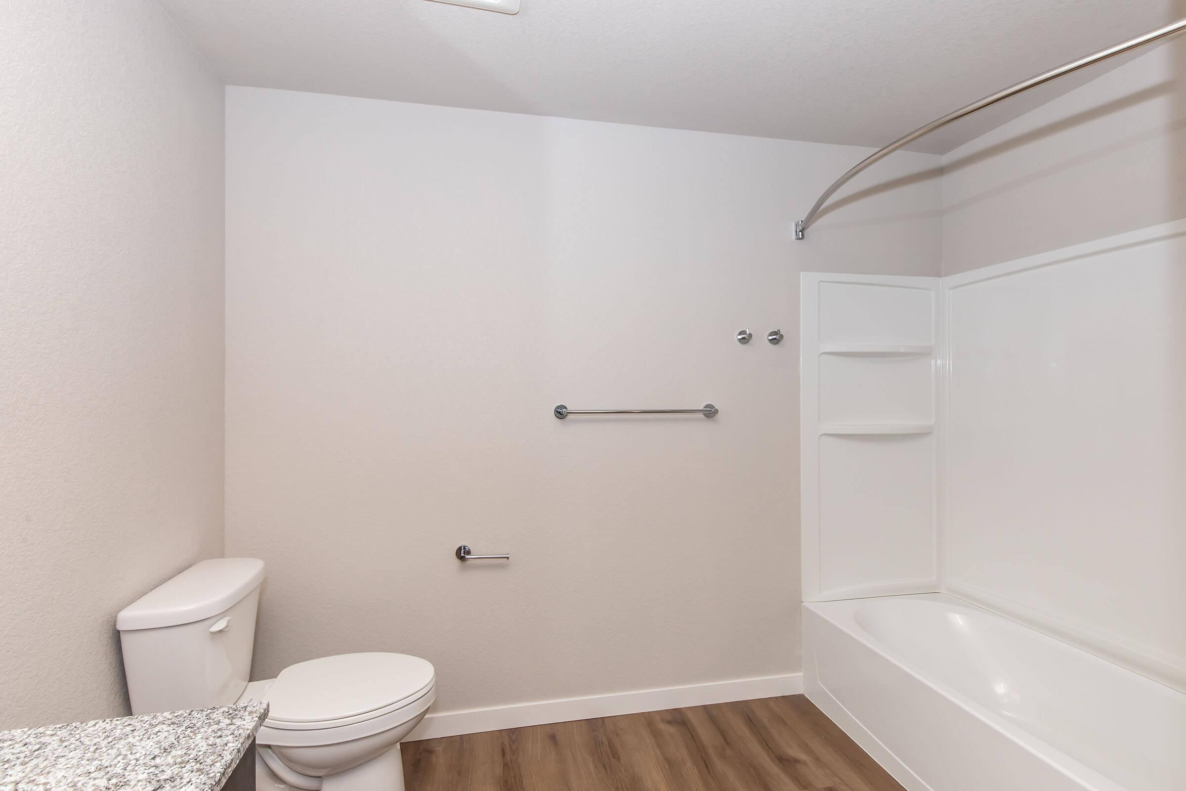 A clean and modern bathroom featuring a white bathtub with a shower curtain rod, a toilet, a wall-mounted towel bar, and a sleek countertop. The walls are painted light gray, and the flooring is made of wood-like laminate. The space is well-lit and minimalist in design.