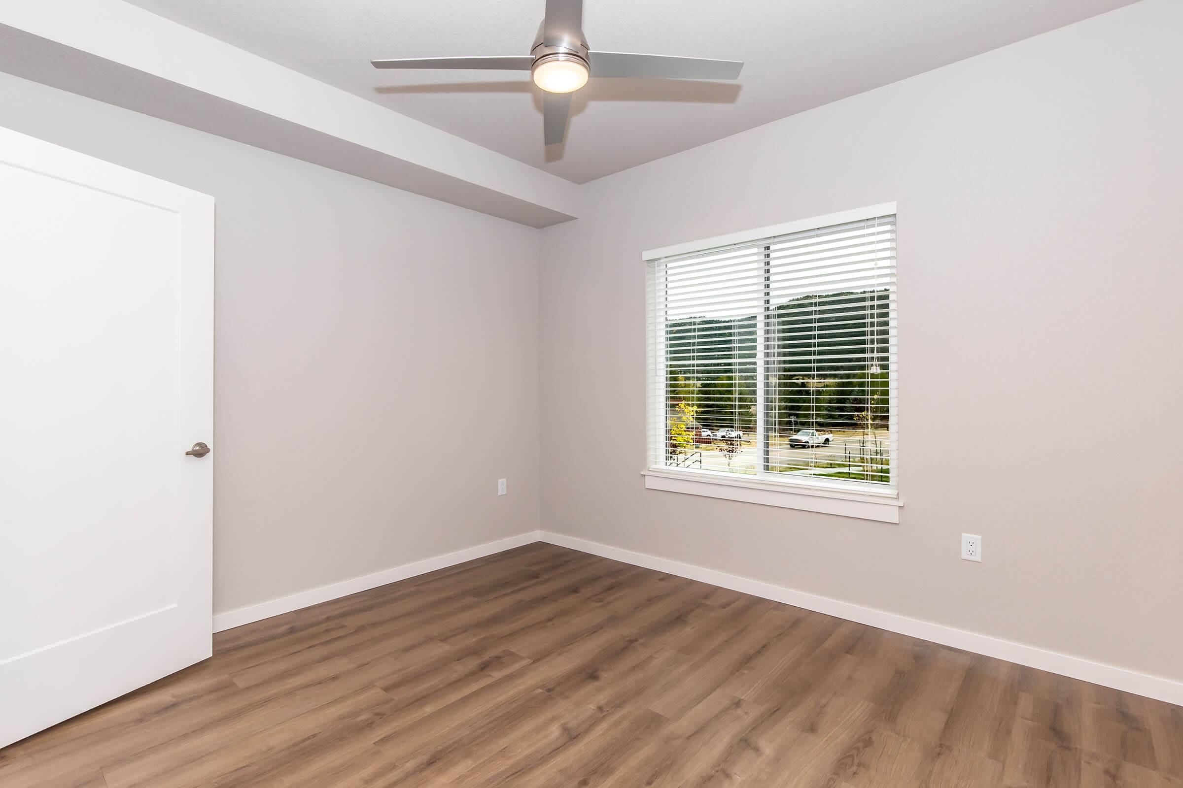 A light gray room with a wooden floor, featuring a window with white blinds, a ceiling fan, and a white door. The walls are bare, giving the space a minimalist look, and there are scenic views visible through the window.