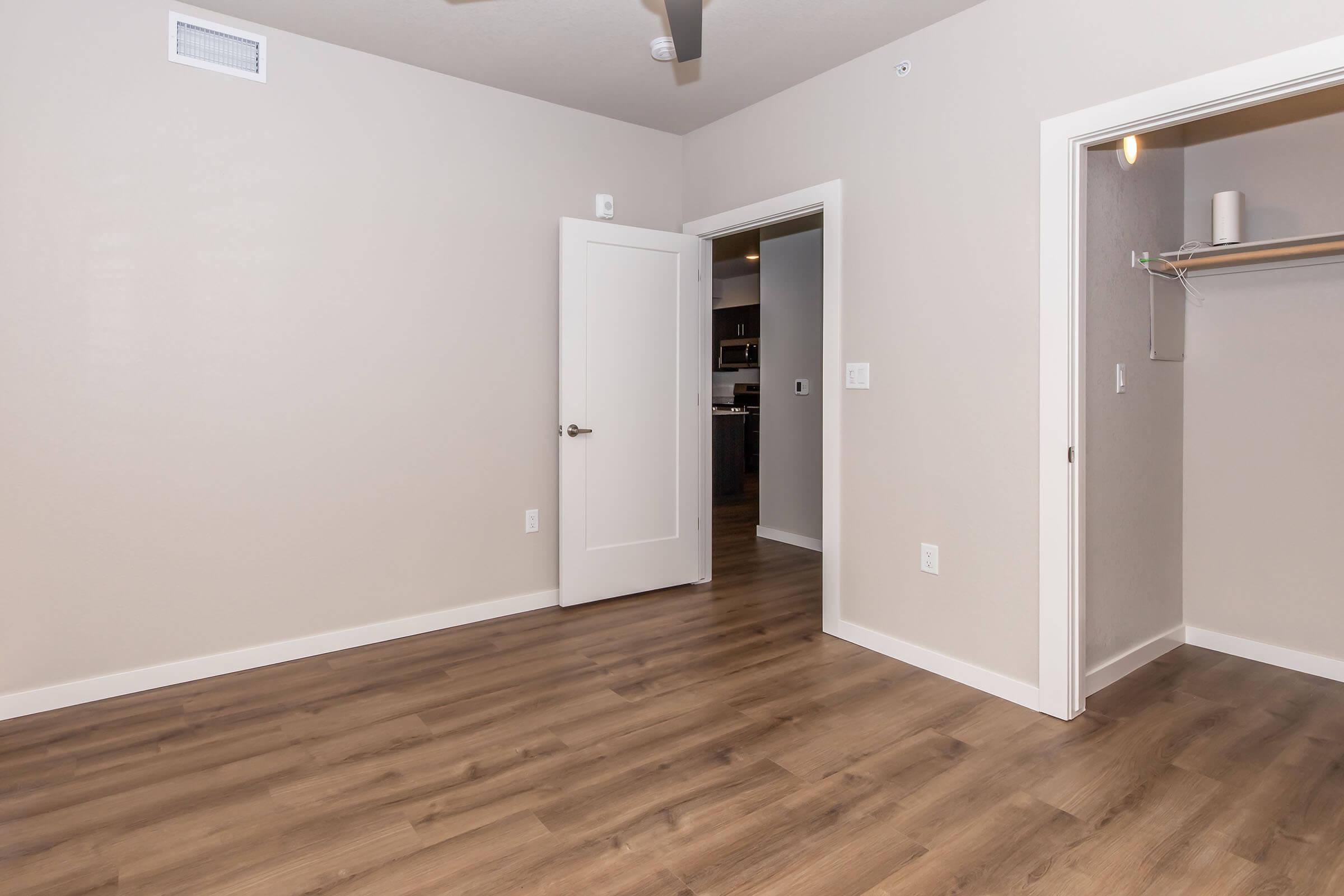 A modern room featuring light-colored walls and wooden flooring. There is a white door slightly ajar leading to another space, and an open closet with a shelf visible. The room is well-lit and simple, with a ceiling fan present. The overall aesthetic is clean and contemporary.