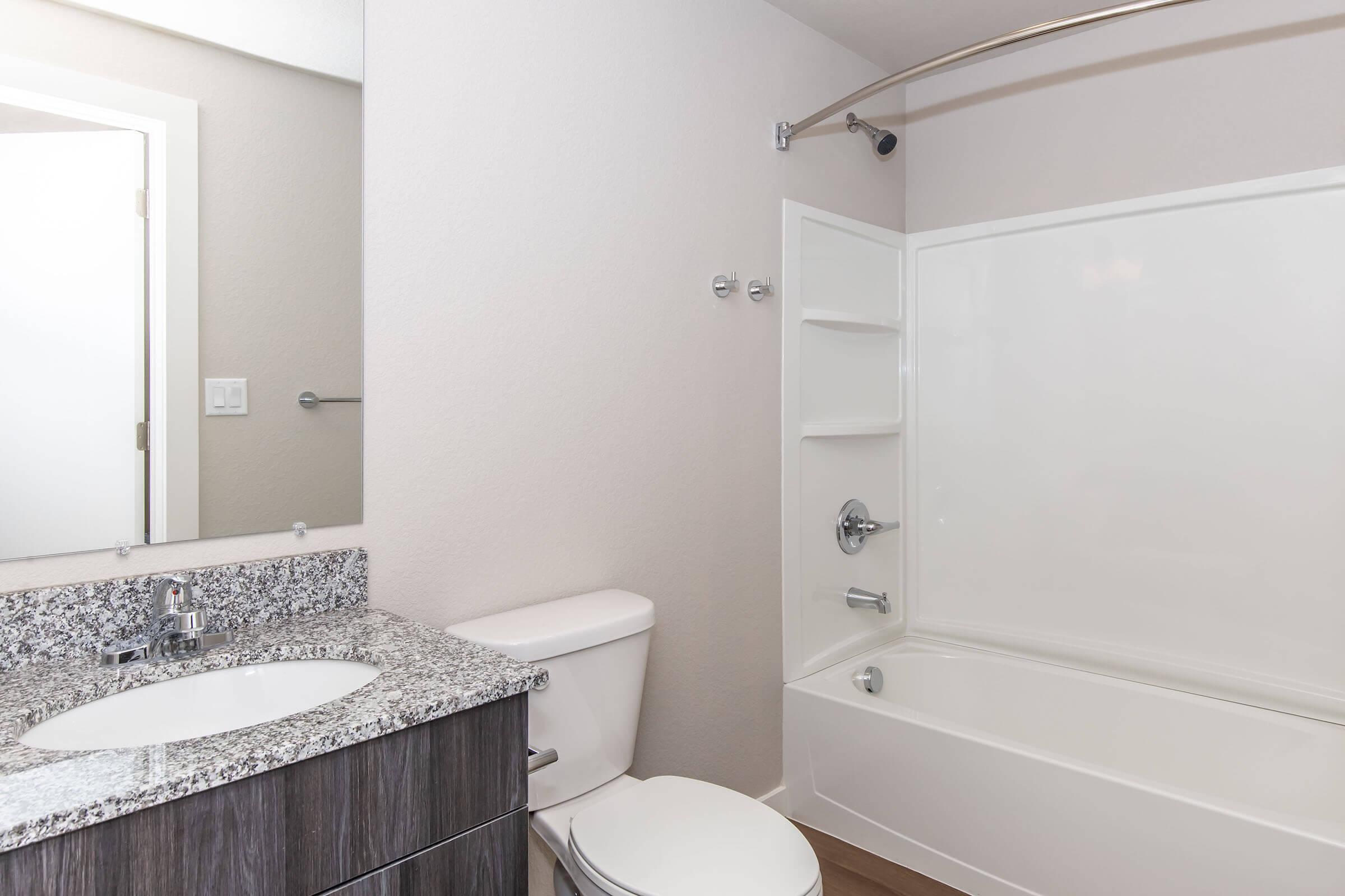 A modern bathroom featuring a marble countertop sink, toilet, and a white bathtub with a shower. The walls are painted in a light color, and there is a mirror above the sink. A shower curtain rod arcs over the bathtub, and the flooring appears to be wood.