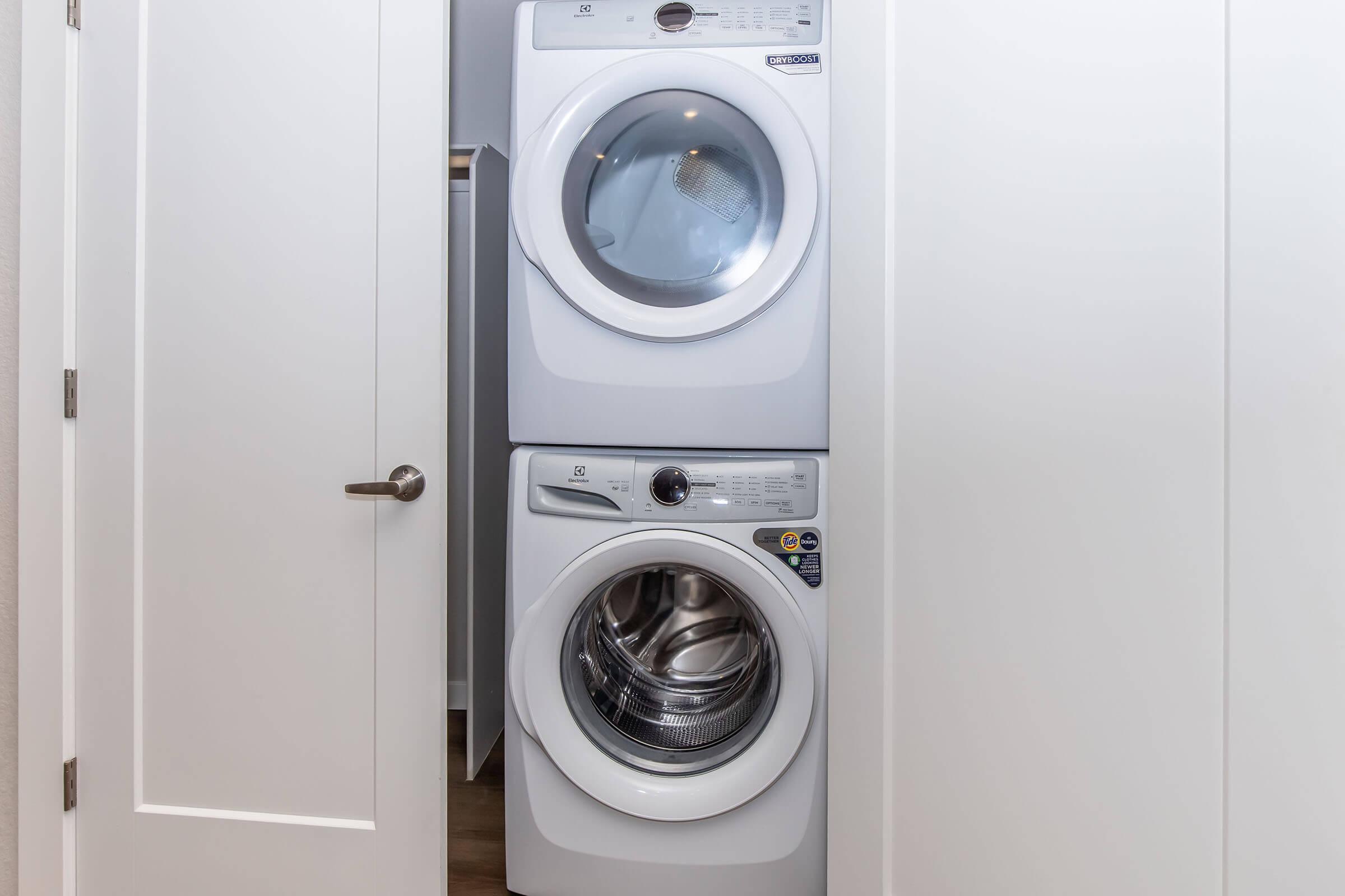 Stacked washing machine and dryer in a white laundry room. The appliances have a modern design with transparent doors, set against a clean white wall and door frame. The space appears tidy and functional, suitable for home laundry tasks.