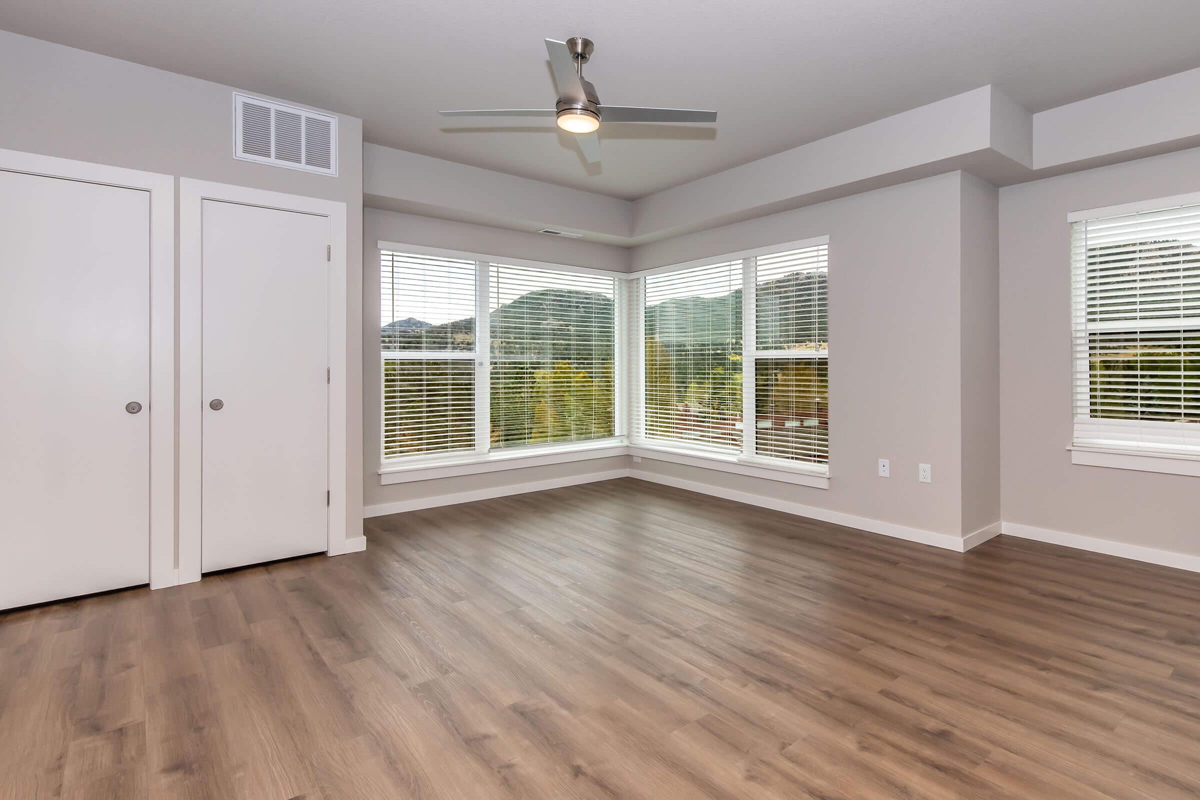 Bright and spacious interior of a modern room with large window featuring mountain views. The room has wood-like flooring, neutral gray walls, and a ceiling fan. Double closet doors are visible, and there are multiple windows with blinds, allowing natural light to enter the space.
