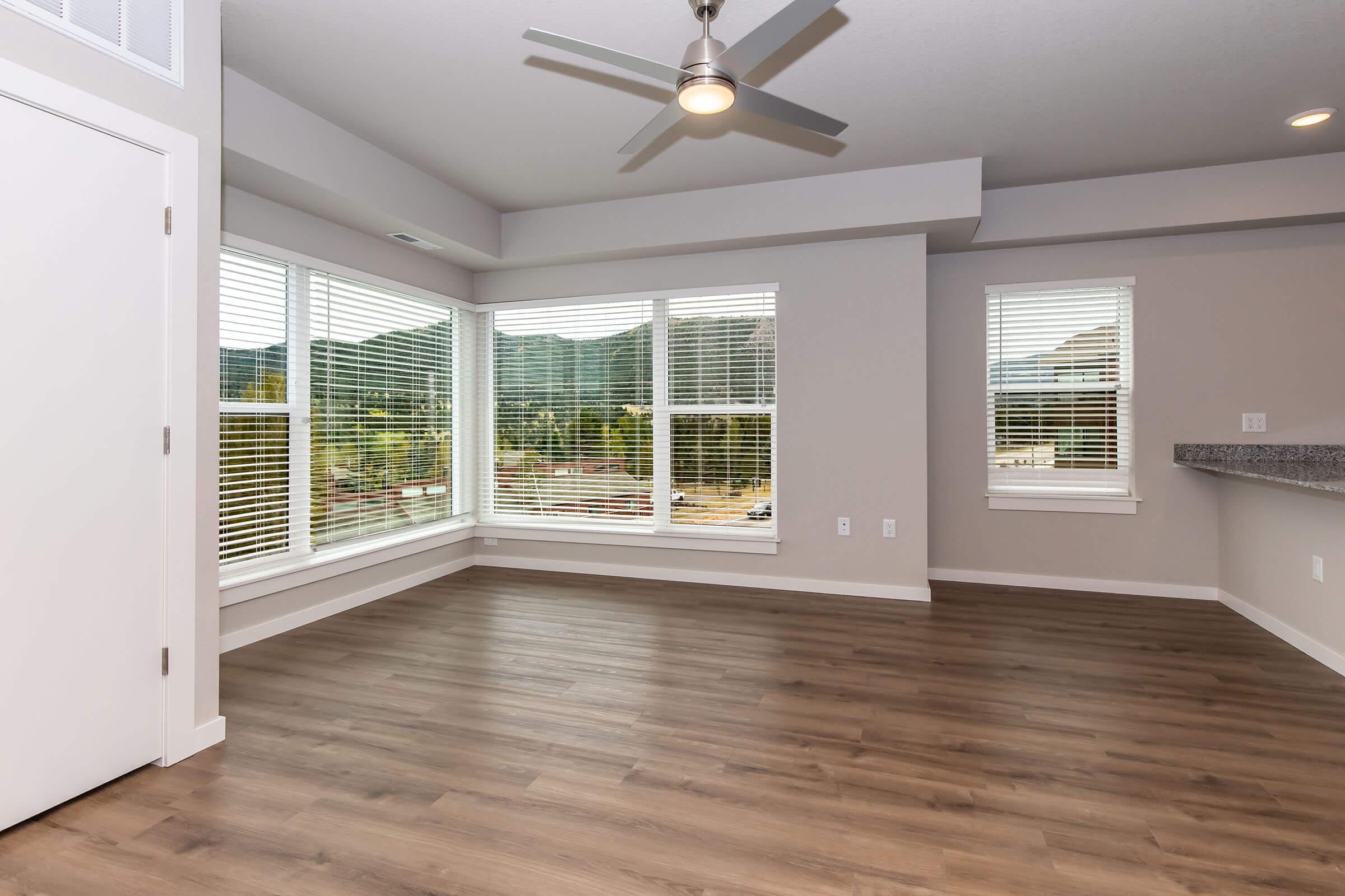 A spacious, well-lit room with large windows featuring white blinds. The floor is made of light-colored wood, and a modern ceiling fan is visible. There's a countertop in the background, suggesting an open layout leading to a kitchen area. The walls are painted in a neutral color, creating a bright atmosphere.