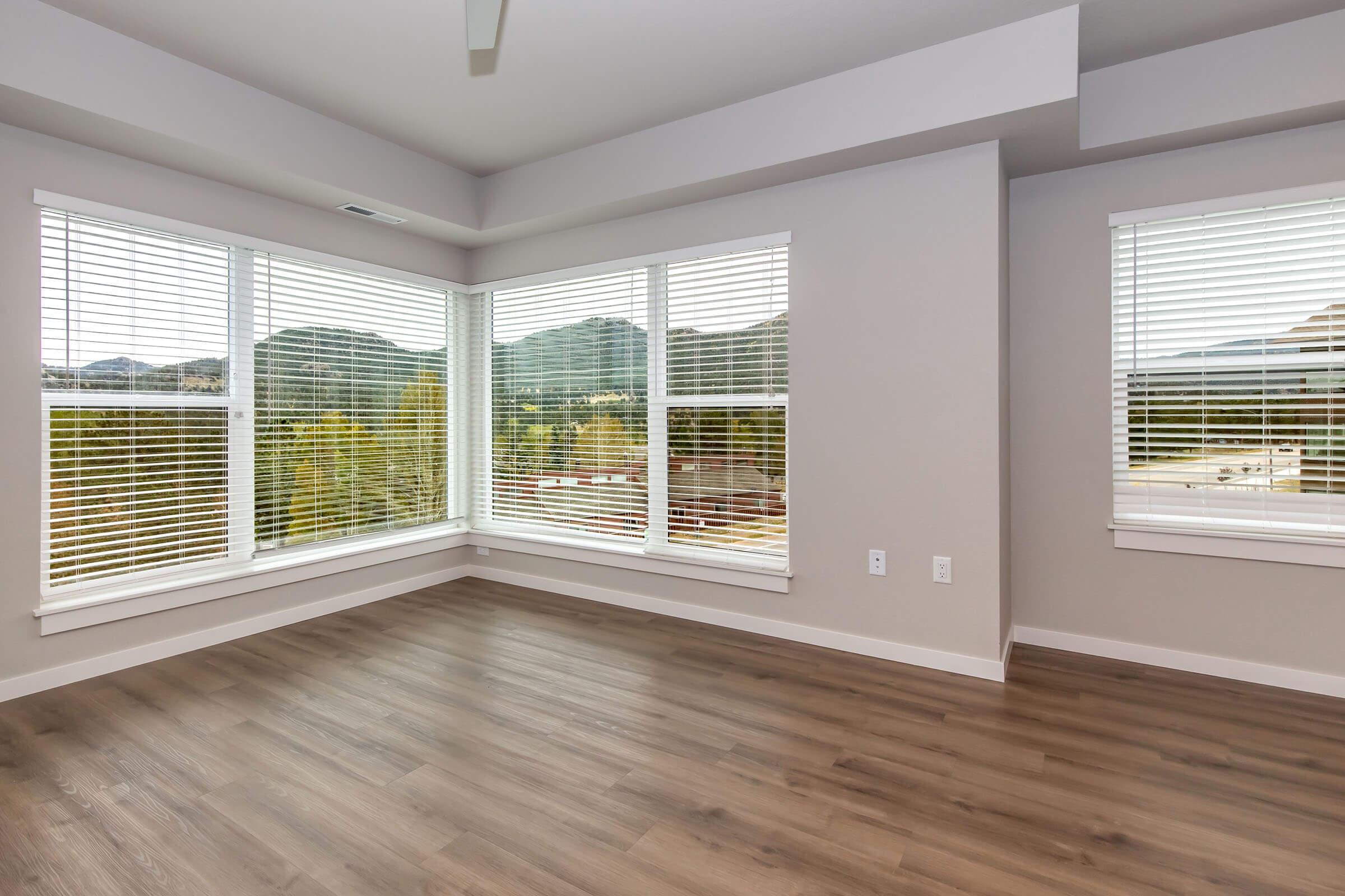 A bright, modern room with large windows featuring wooden blinds. The floor is light-colored wood, and the walls are painted in a soft, neutral tone. Outside the windows, green mountains and trees can be seen, creating a scenic view. Natural light fills the space, emphasizing its airy feel.