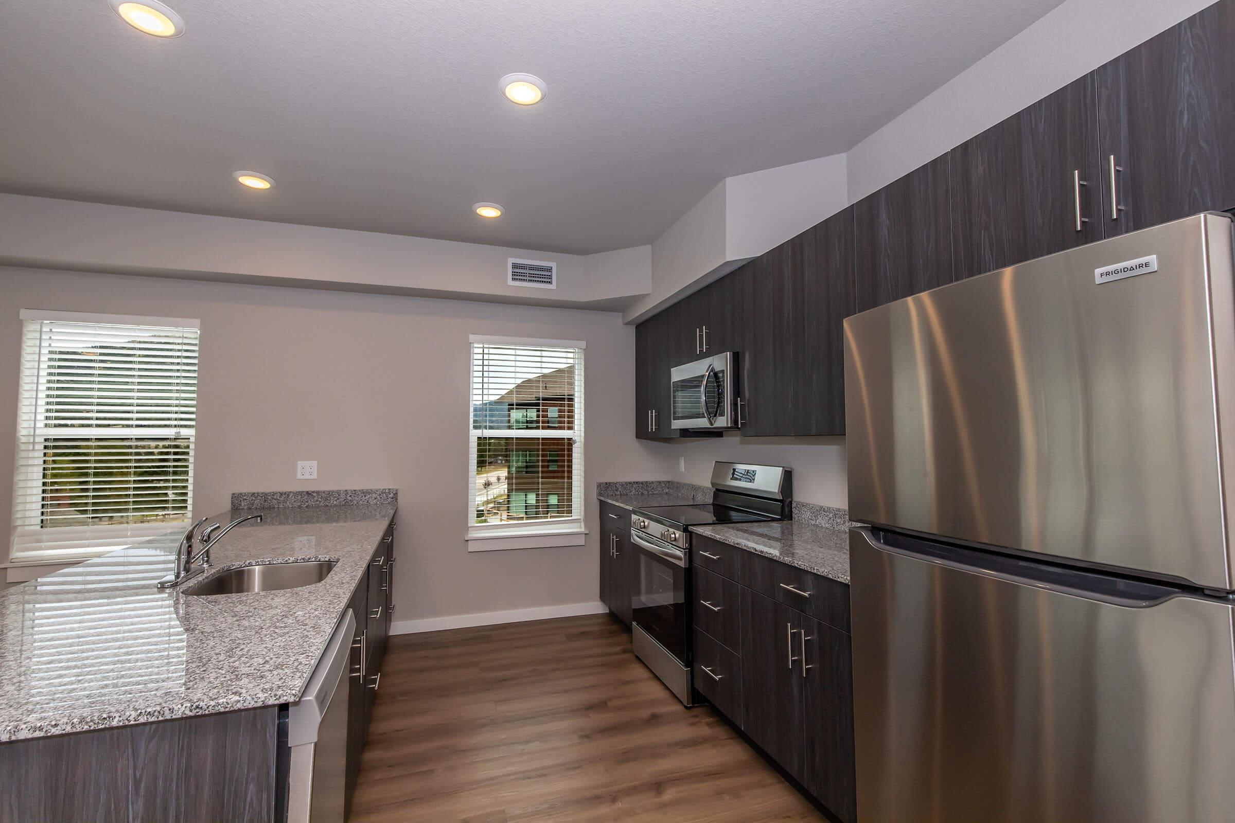 Modern kitchen featuring dark wood cabinetry, granite countertops, a stainless steel refrigerator, and an oven. Large windows provide natural light, and the open layout includes a kitchen island with a sink. The flooring is a light wood finish, enhancing the contemporary feel of the space.