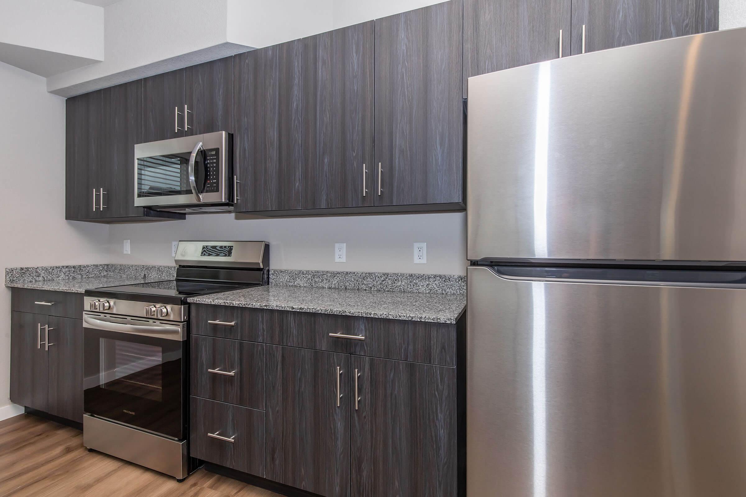 Modern kitchen with dark wood cabinets, stainless steel appliances including a microwave, oven, and refrigerator. The countertop is made of light-colored granite, and the flooring is wood-like. The overall design is sleek and contemporary, featuring a minimalist aesthetic.