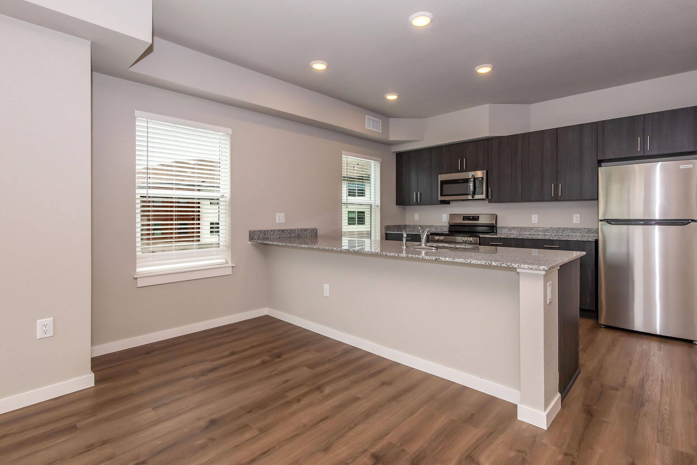 Modern kitchen featuring dark wood cabinetry, stainless steel appliances, and a granite countertop. The space includes a window with blinds and has hardwood-like flooring. Bright recessed lighting enhances the contemporary look, with an open layout that connects to the living area.