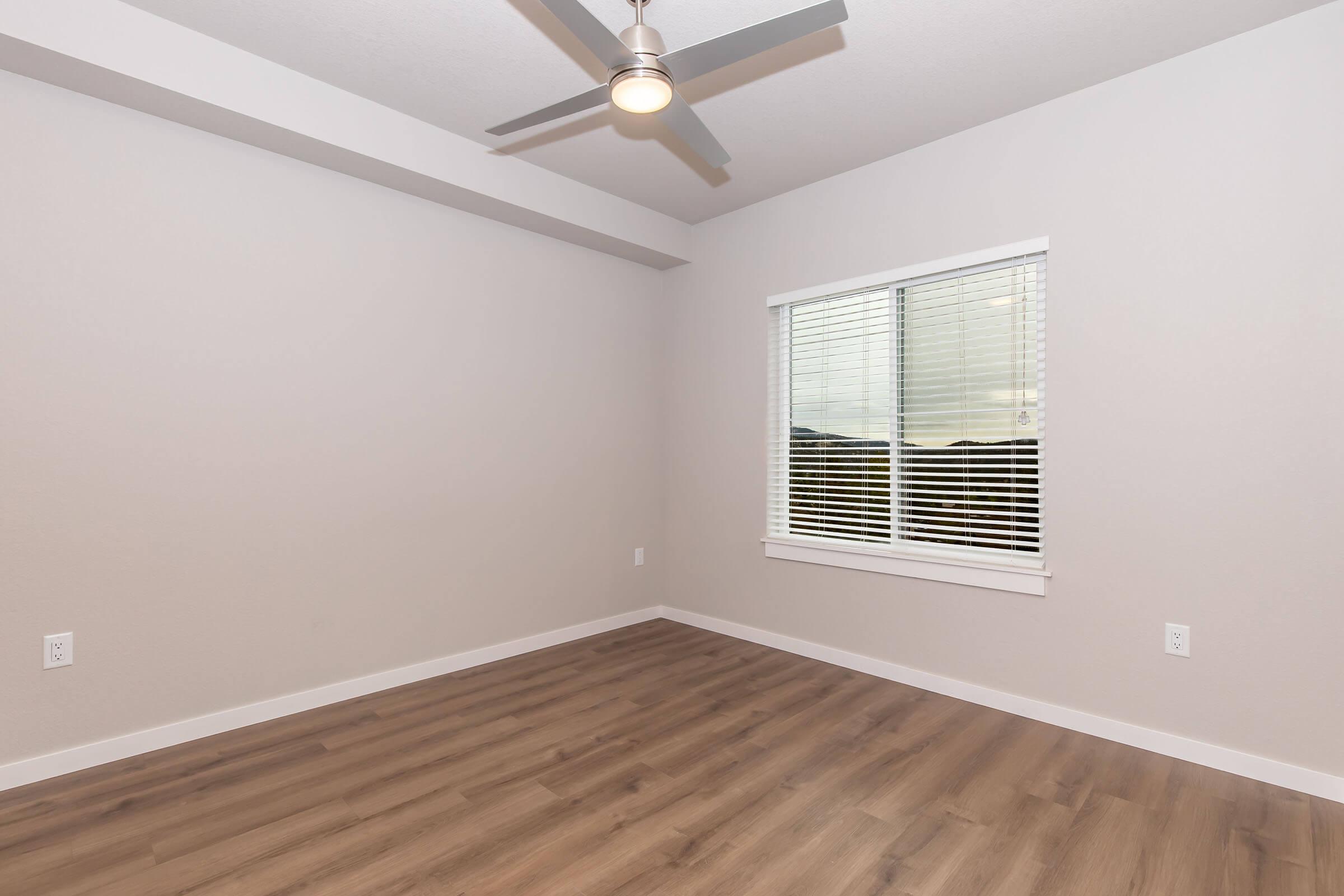 Empty room with light gray walls and a ceiling fan. A window with white blinds allows natural light, and the floor is covered in light wood laminate. The room has a spacious feel, with no furniture present, emphasizing the clean and minimalistic design.