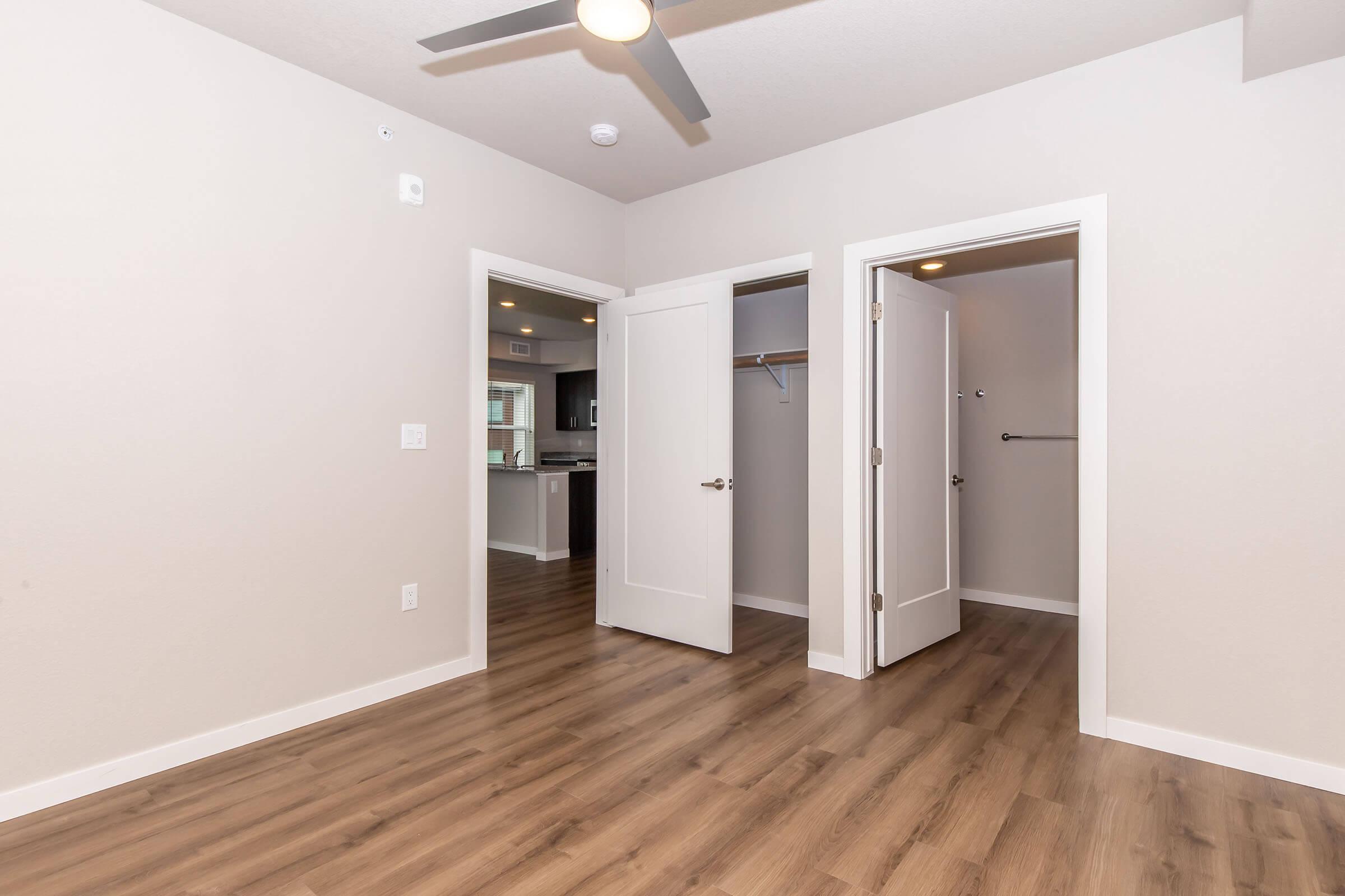 A well-lit room featuring light-colored walls and wooden flooring. There are two doorways leading to other spaces, one of which is a closet. A ceiling fan is installed, and the background hints at a modern kitchen area visible through the open door.