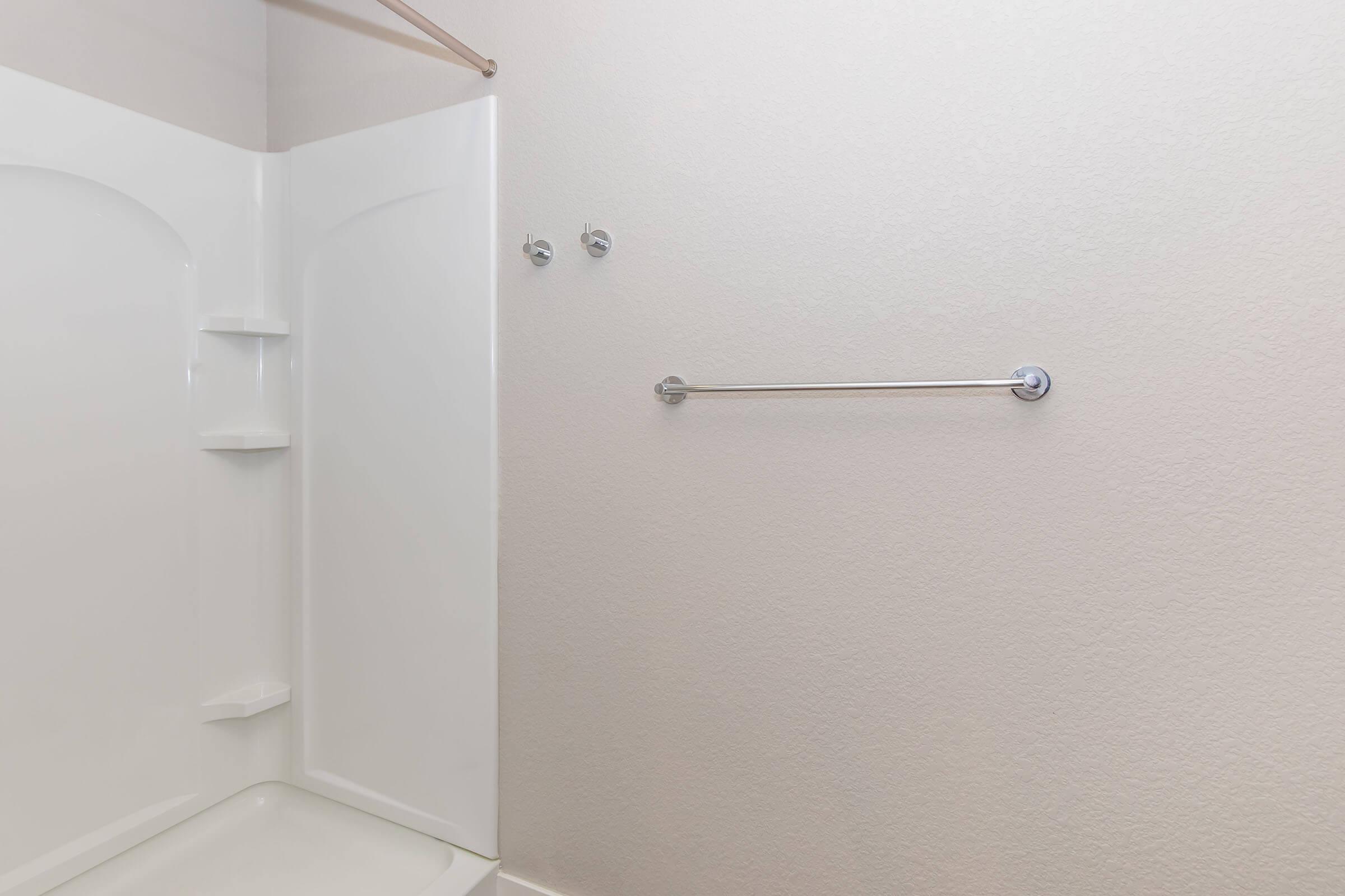 A clean and empty bathroom featuring a white bathtub and shower unit, with two wall-mounted hooks and a chrome towel bar. The walls have a light texture, and the overall space is bright and uncluttered.