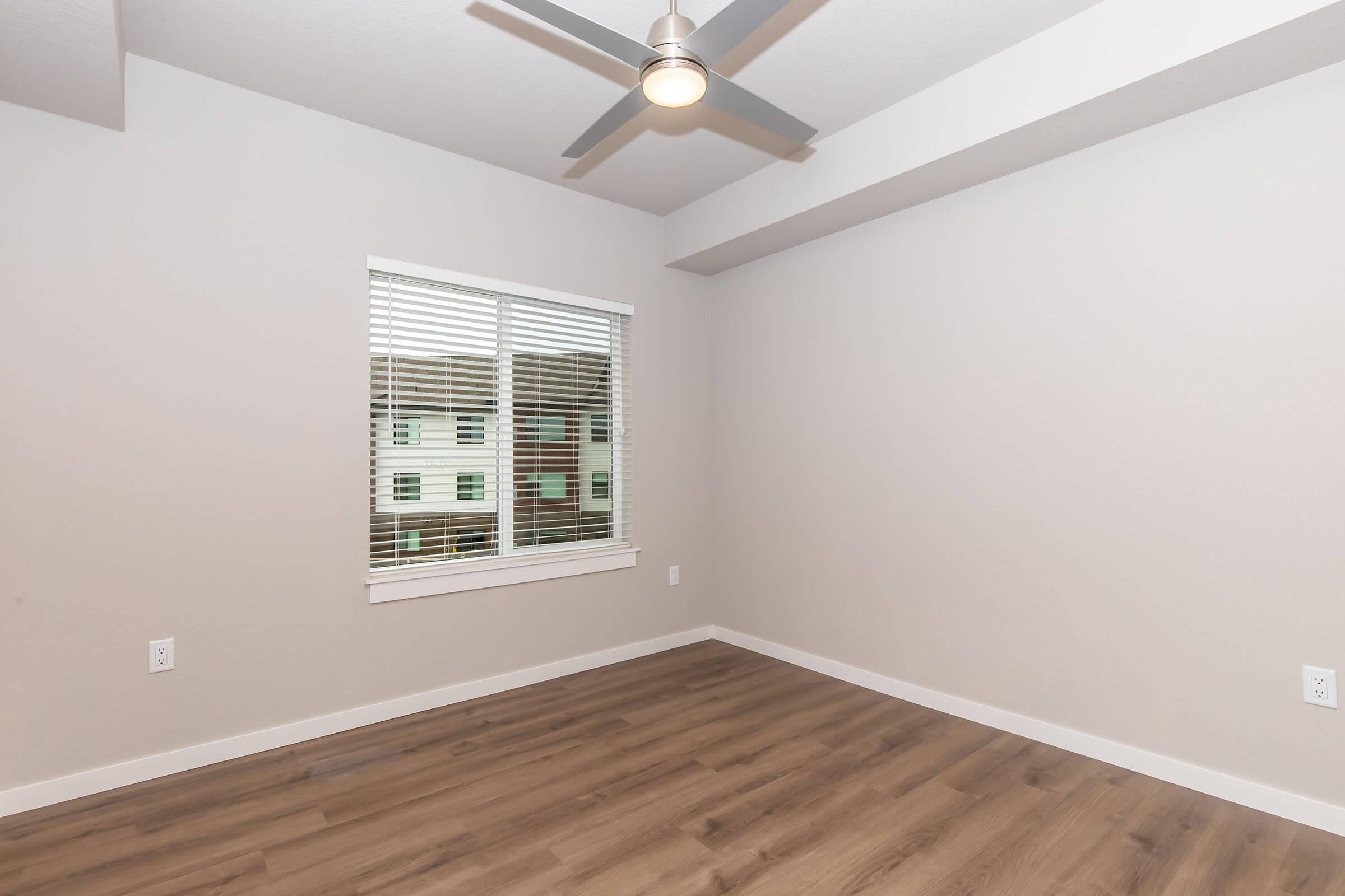 Empty room with light gray walls and a ceiling fan. There is a window with white blinds, showing an exterior view. The floor is covered in wooden planks, creating a warm and natural atmosphere. The space appears clean and freshly painted, suitable for new furnishings.