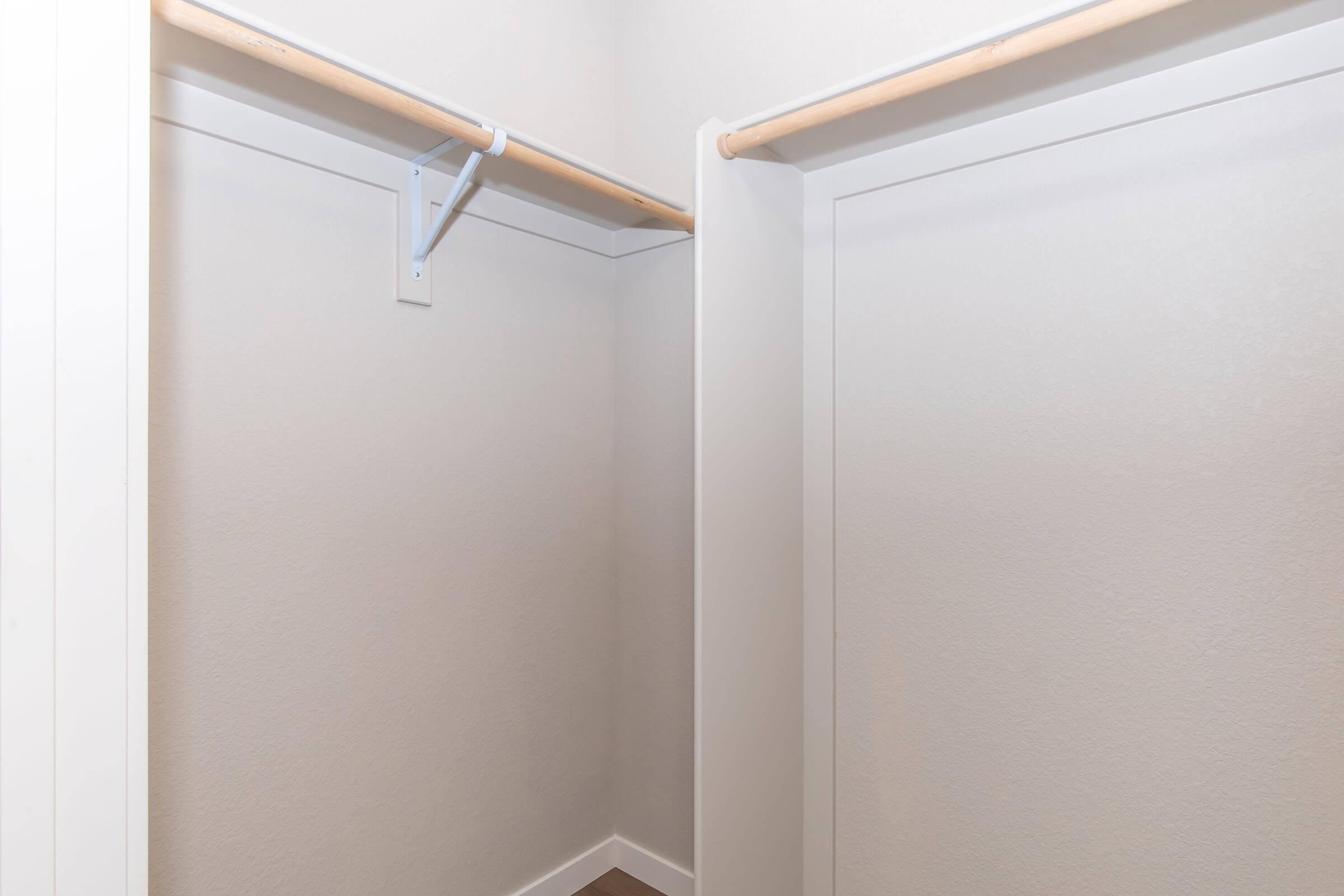 Two empty closet spaces with light-colored walls and a wooden floor. Each closet features a horizontal wooden rail for hanging clothes, with clean lines and a minimalist design. The lighting is soft, creating a simple and uncluttered atmosphere.