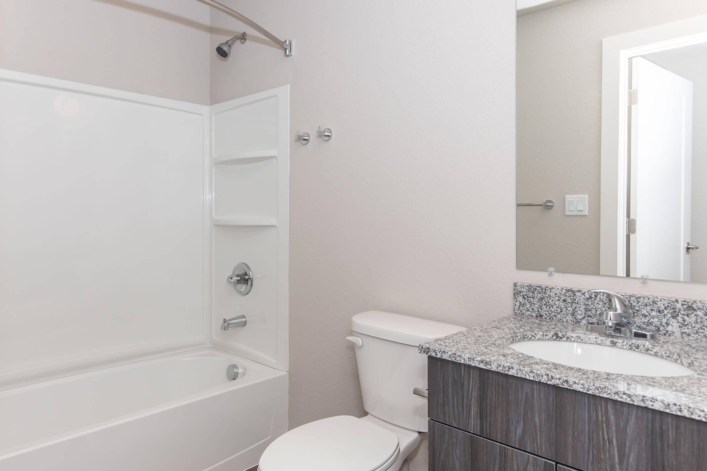 A clean, modern bathroom featuring a white bathtub with a shower, a gray granite countertop sink, a toilet, and a large mirror. Light-colored walls and a door are visible, creating a simple and uncluttered aesthetic.
