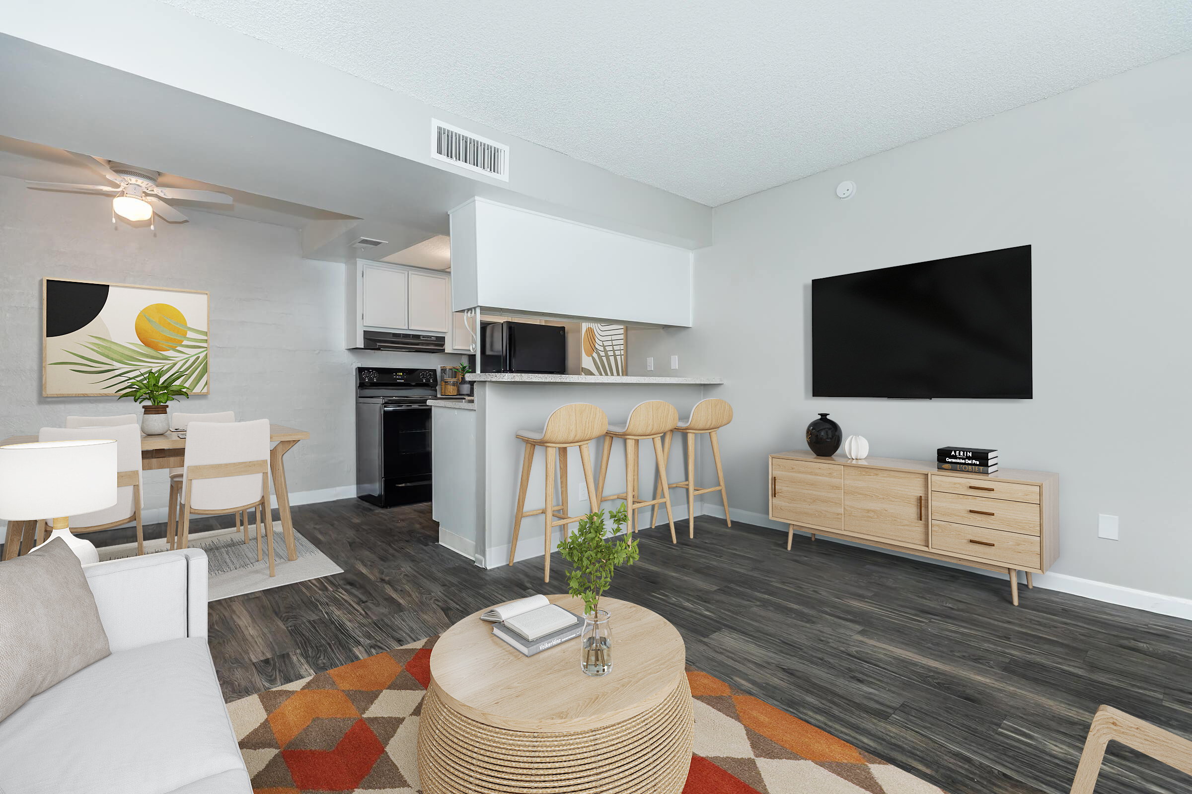 Modern living room and kitchen area featuring a beige sofa, circular coffee table, and a dining set. The kitchen has black appliances and light cabinets, while a large TV is mounted on the wall. A vibrant wall art piece and a decorative plant add color and style to the space.