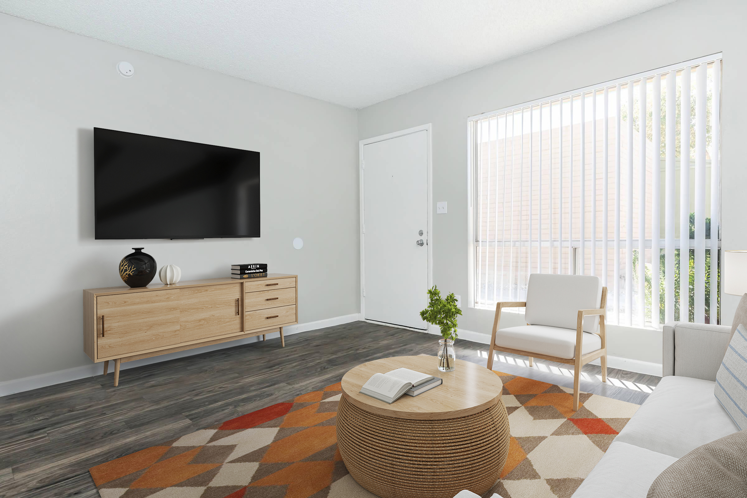 A modern living room featuring a flat-screen TV mounted on a light gray wall. There's a wooden TV cabinet, a round coffee table with a book on it, and a beige armchair. A large window with vertical blinds allows natural light in, and a colorful geometric rug adds a touch of warmth to the space.