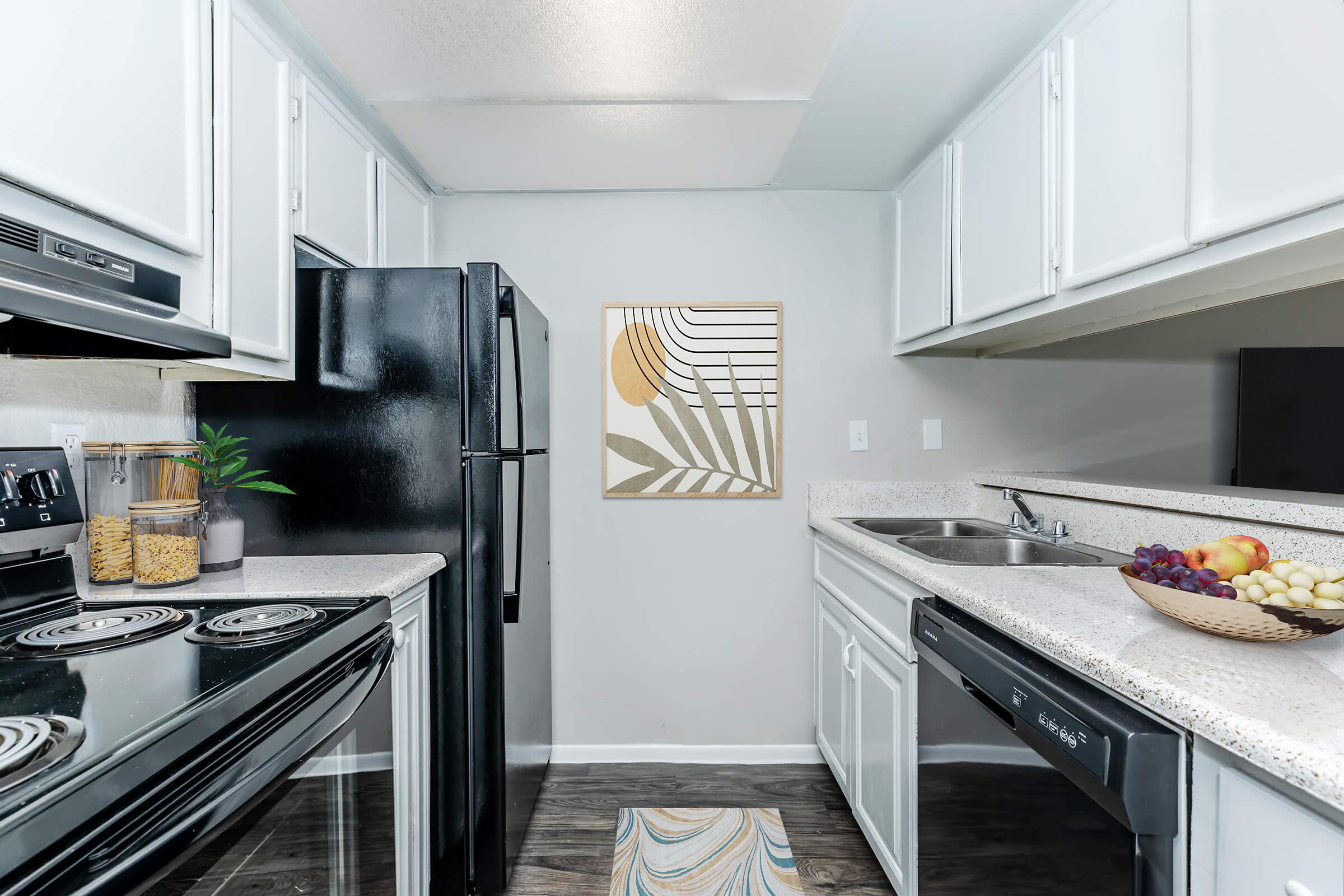 A modern kitchen featuring black appliances, white cabinetry, and a light countertop. There is a framed artwork on the wall, a bowl of assorted fruits on the counter, and a small plant near the sink, creating a fresh and inviting atmosphere.