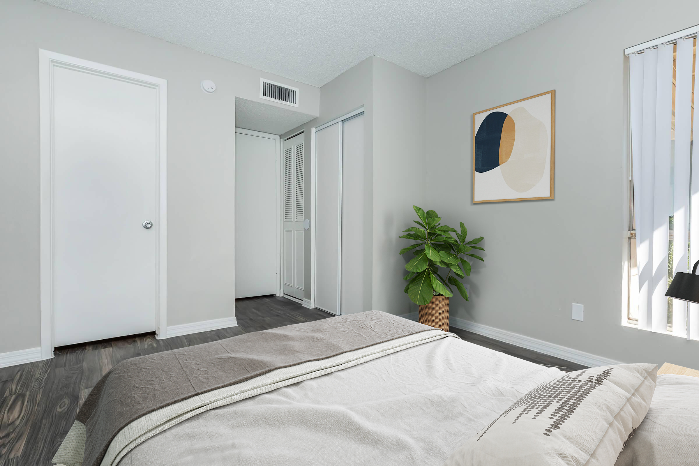 A well-lit bedroom featuring a neatly made bed with a beige and gray coverlet. There is a green potted plant beside the bed, and a doorway leading to a closet area can be seen. The walls are painted light gray, and a piece of modern art hangs on the wall. Large windows allow natural light to enter the space.