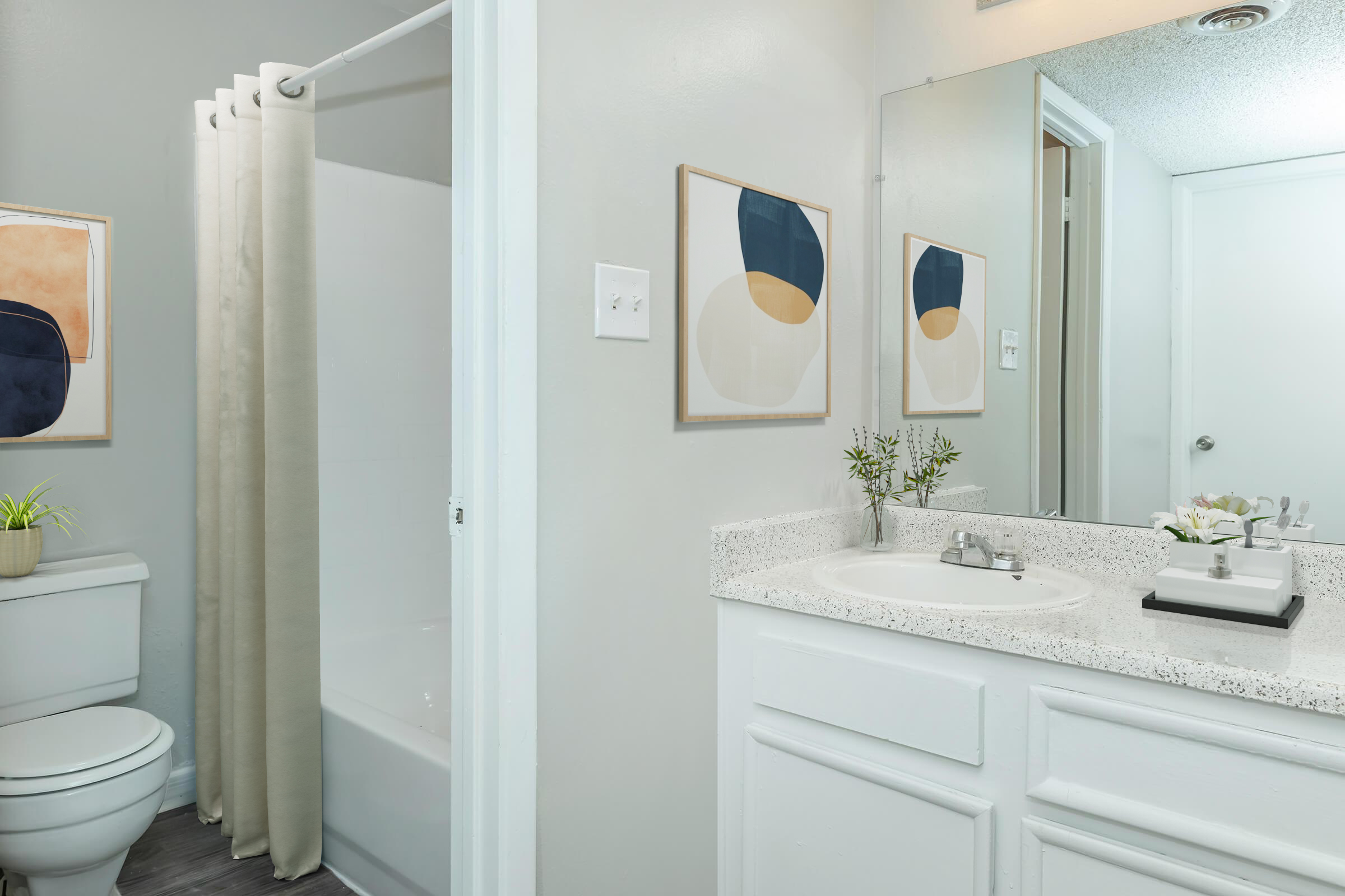 Brightly lit bathroom featuring a white sink with a counter, a shower area with a beige curtain, and light grey walls. Two framed abstract art pieces hang on the wall, and a small plant decorates the counter. A toilet is visible, adding to the clean and modern design of the space.