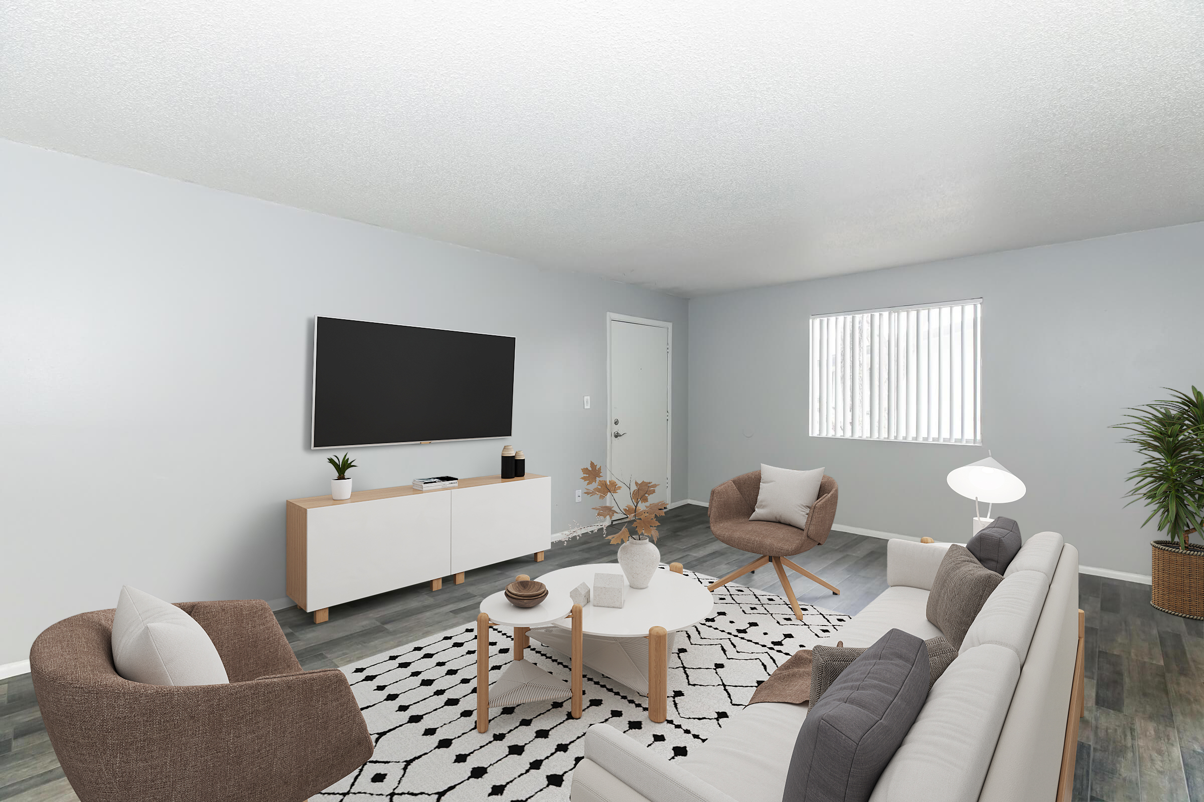 A modern living room featuring a light gray wall, a flat-screen TV mounted on the wall, a white sideboard, a cozy sofa with pillows, two stylish armchairs, a round coffee table with decorative items, and a plant. Natural light enters through a window with blinds, illuminating the space.