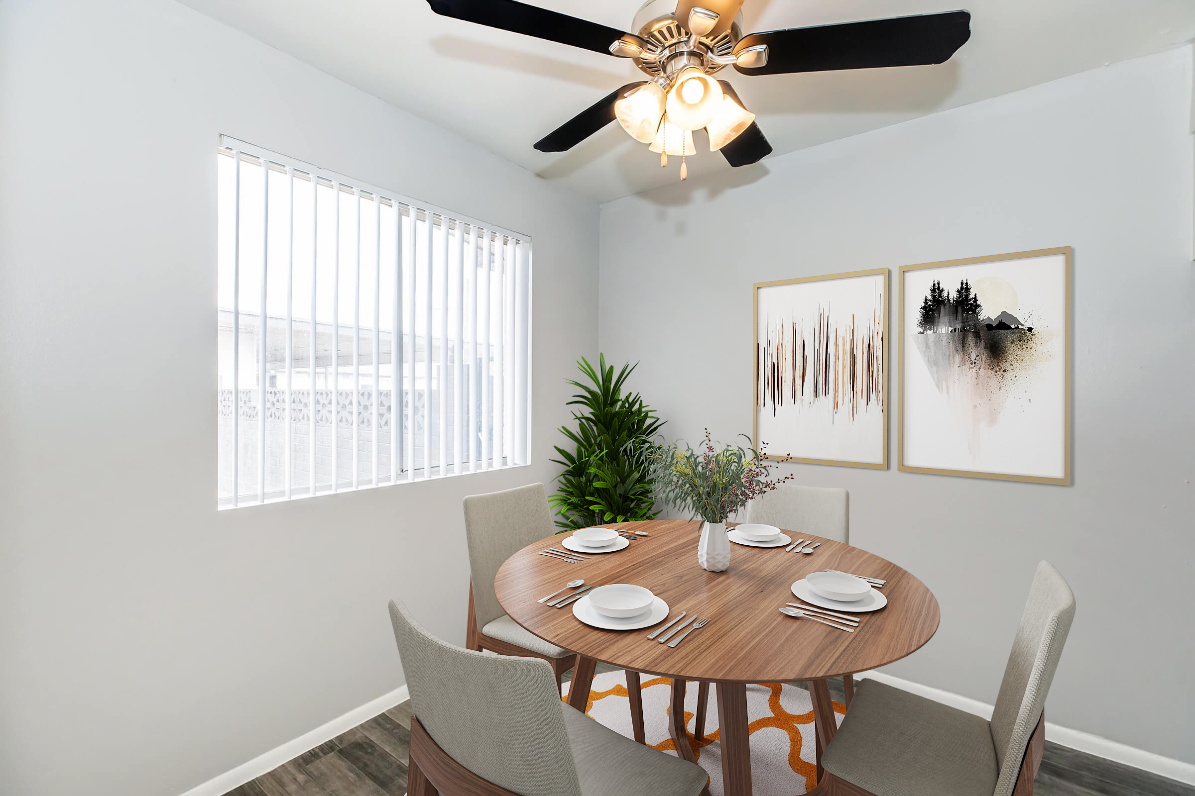 A bright dining area featuring a round wooden table set for four with plates and utensils. The walls are painted light gray, adorned with two framed art pieces. A small plant adds greenery, and a ceiling fan with a light fixture hangs above. Large window with vertical blinds allows natural light to fill the space.