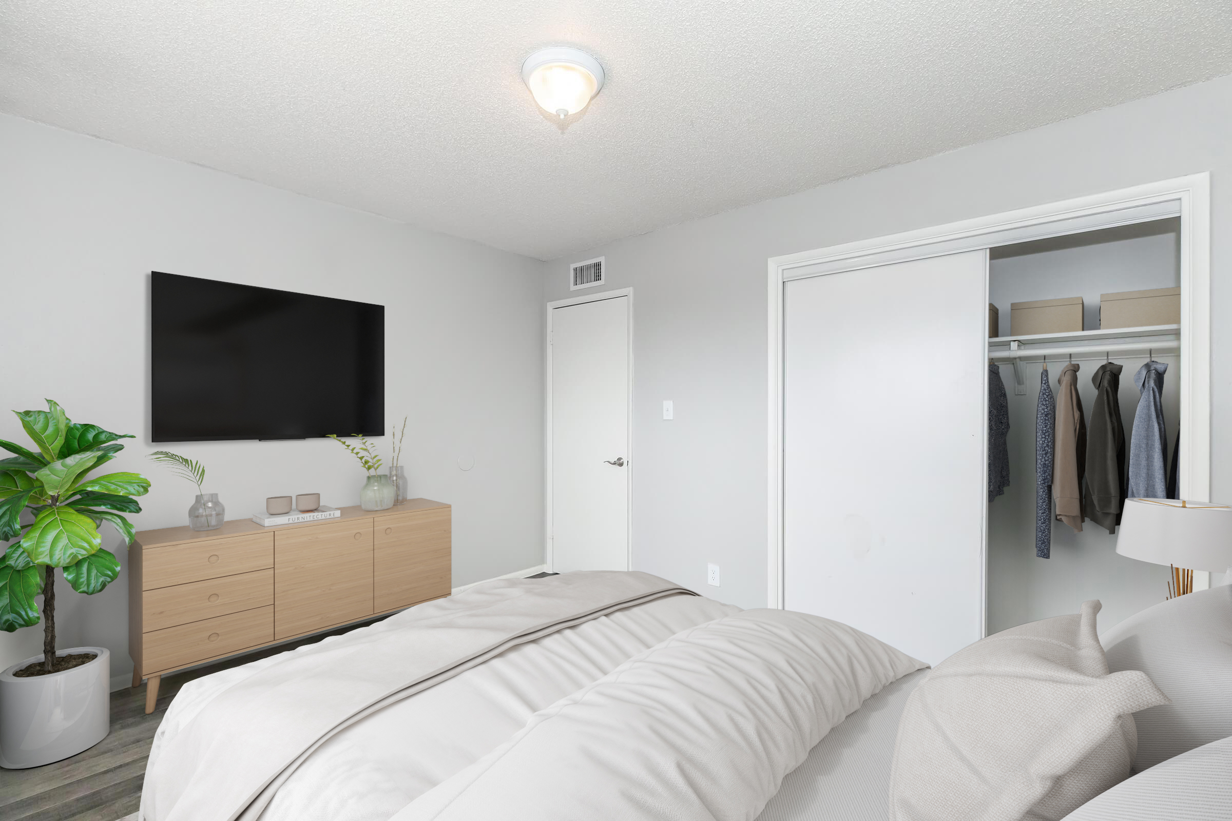 A modern bedroom featuring a large bed with fluffy bedding, a wall-mounted television, and a wooden dresser. A potted plant adds a touch of greenery, and a closet is partially visible with organized clothing. The walls are painted in a light gray tone, contributing to a calm and inviting atmosphere.