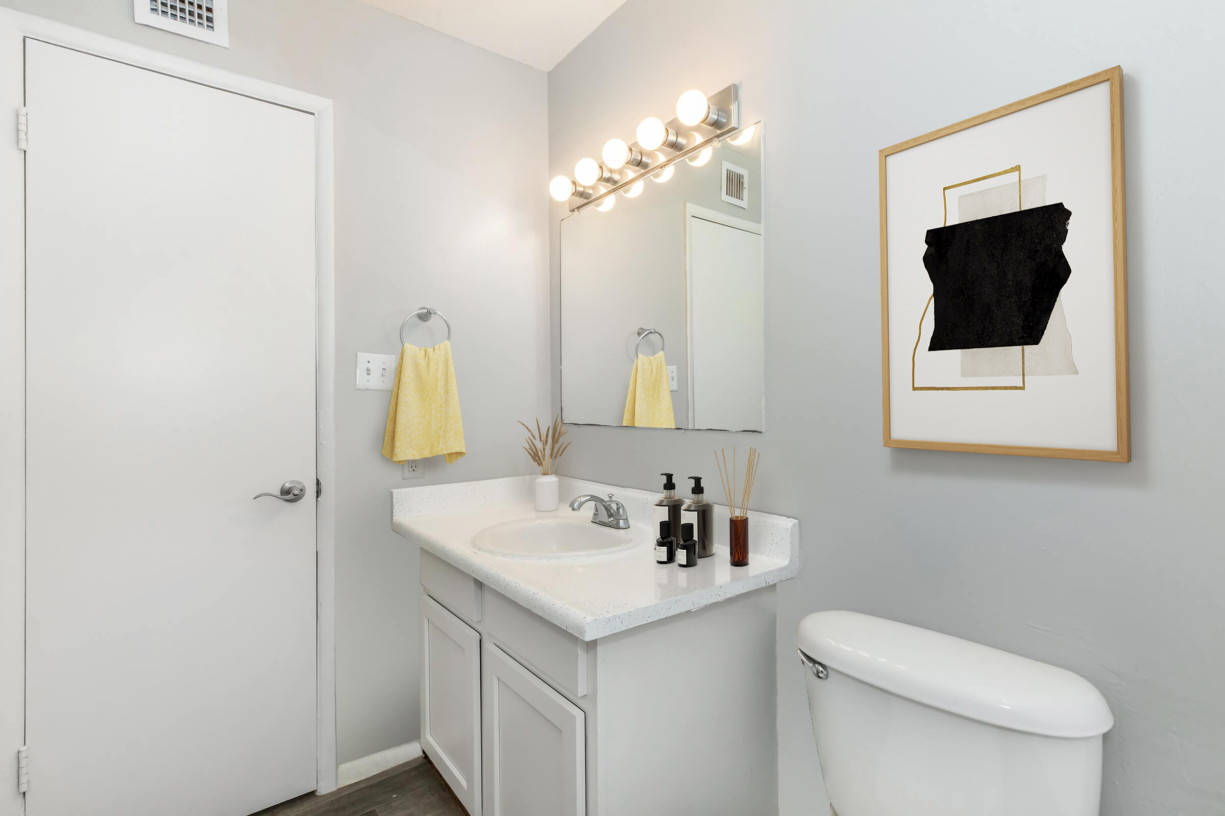 A modern bathroom featuring a white sink with skincare products, a large mirror above it, a yellow hand towel hanging on the side, and an abstract framed artwork on the wall. The decor is minimalist with gray walls and a clean, organized appearance.