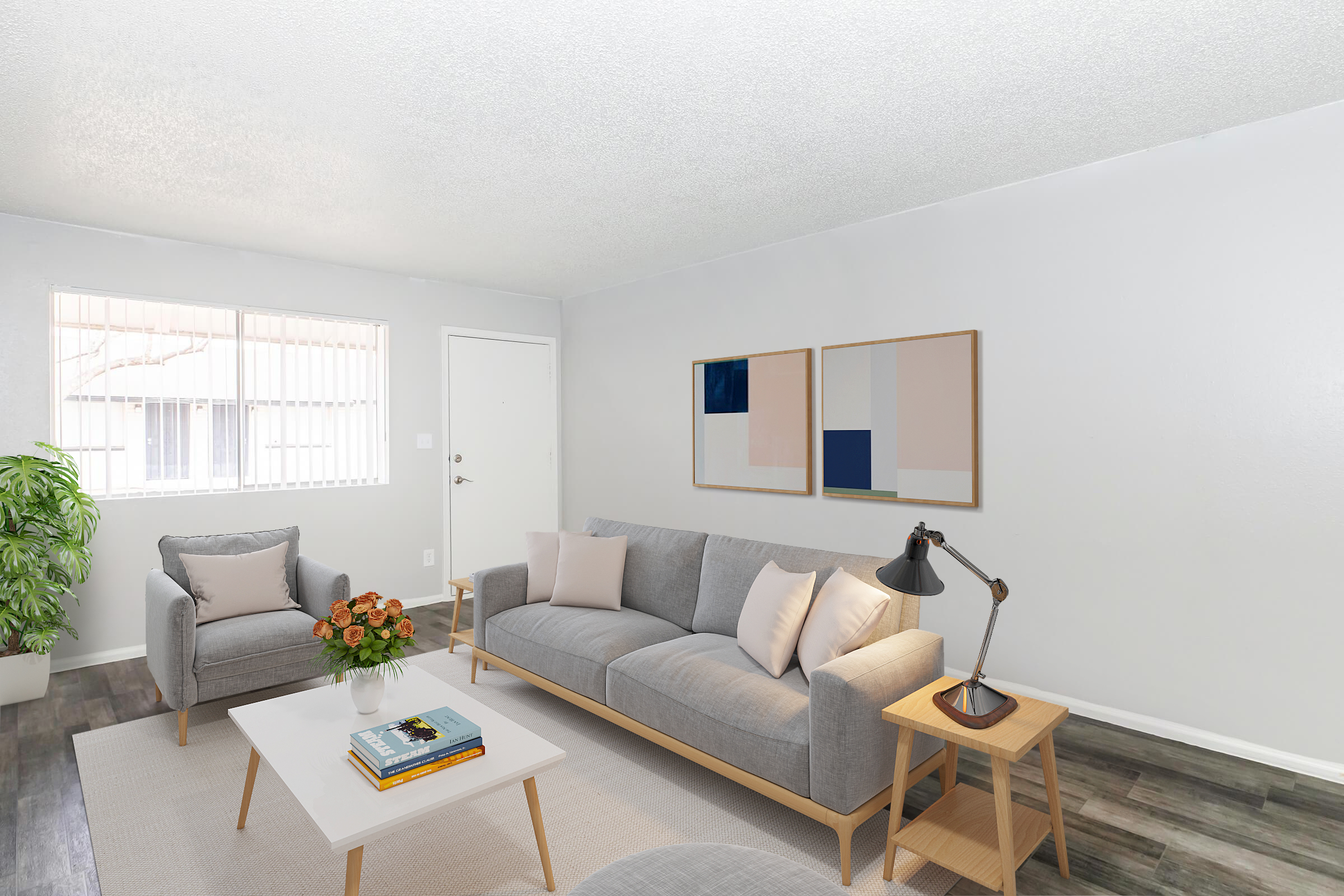 A modern, minimalist living room featuring a light gray sofa with decorative cushions, a small side table with a lamp, and a potted plant. There are two abstract art pieces on the wall, a white coffee table with books, and a cozy chair with flowers nearby. Natural light enters through the window.
