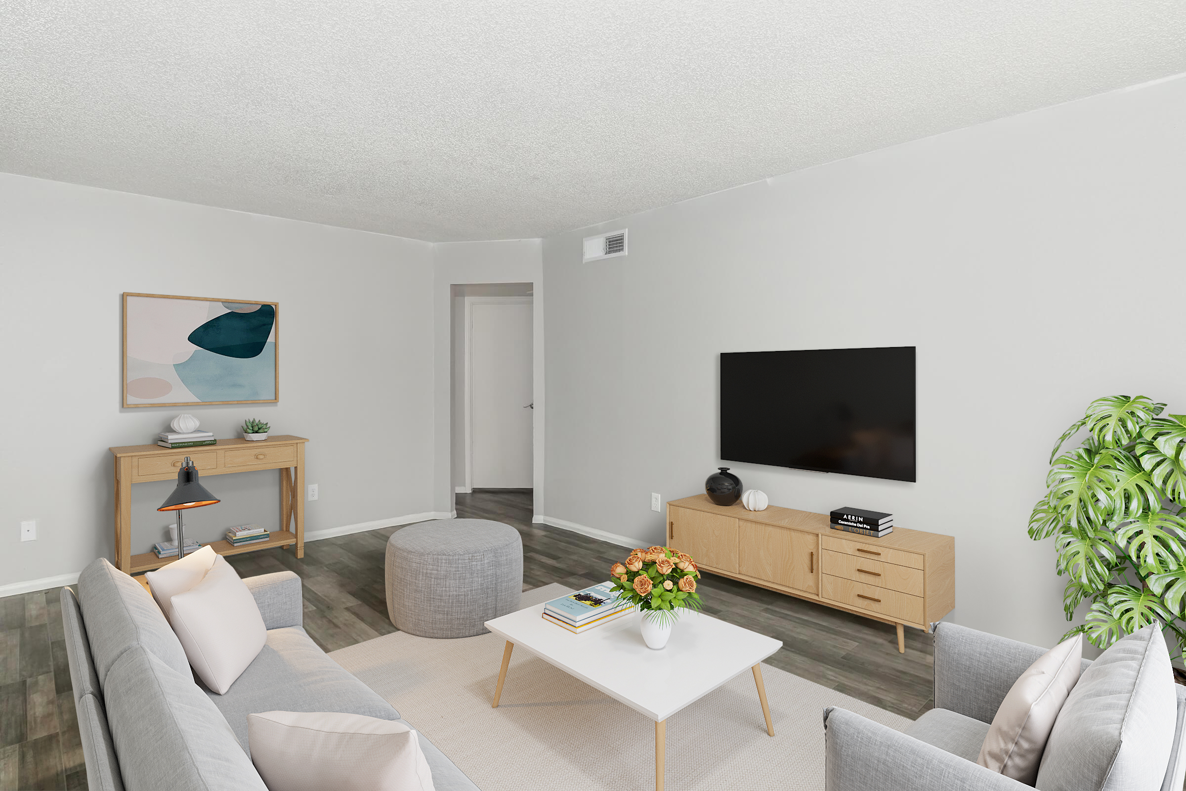 A modern living room featuring a light gray sofa and chairs, a white coffee table, and a wooden TV stand with a flat-screen TV. The walls are painted in a soft gray, and there is a small table with a decorative plant. Natural light fills the space, enhancing the contemporary decor.