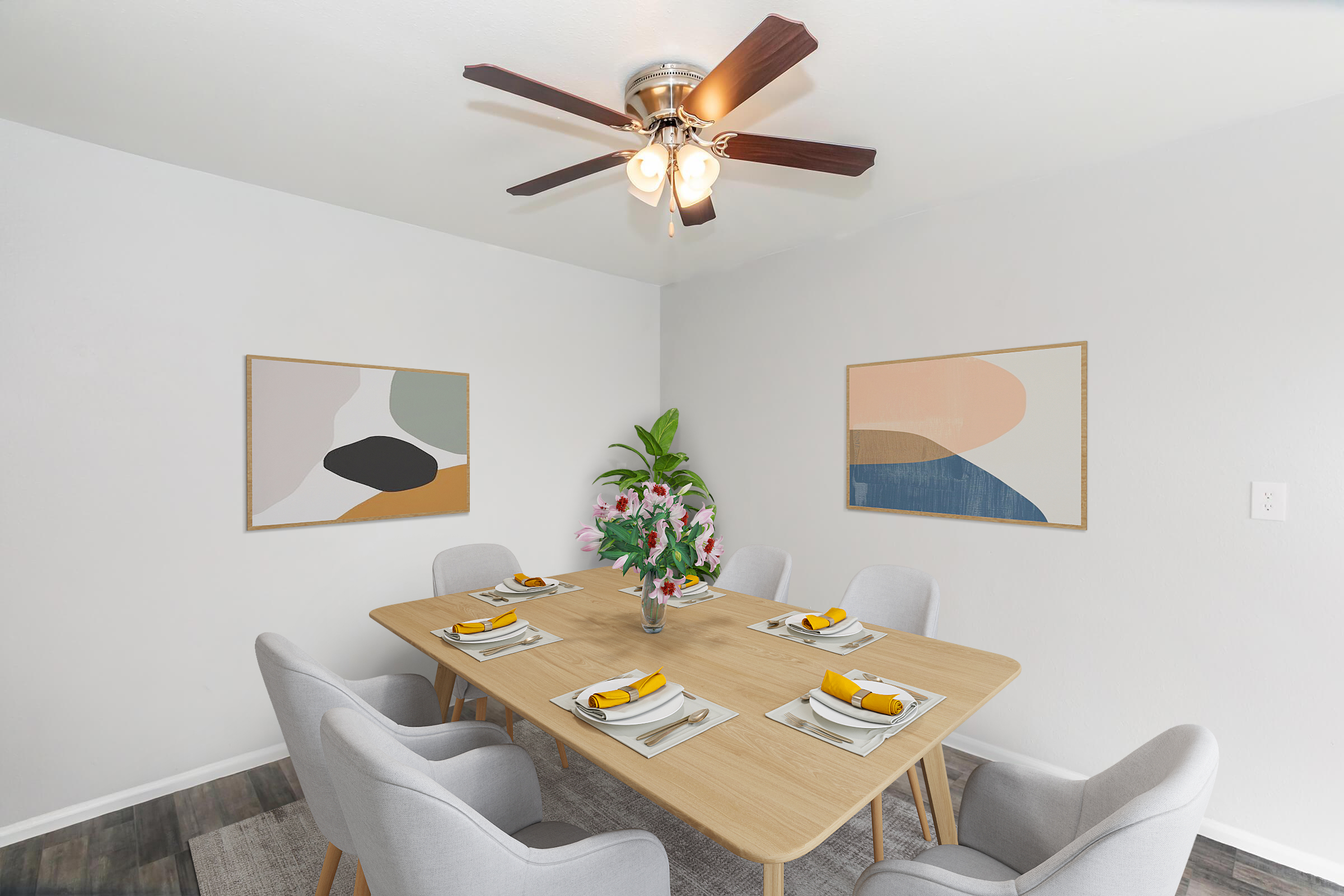 A modern dining room featuring a wooden table set for six with yellow and white dishes. There are gray upholstered chairs, a floral centerpiece, and two abstract wall art pieces. Soft natural light illuminates the space, complemented by a ceiling fan. The walls are painted in a light color.