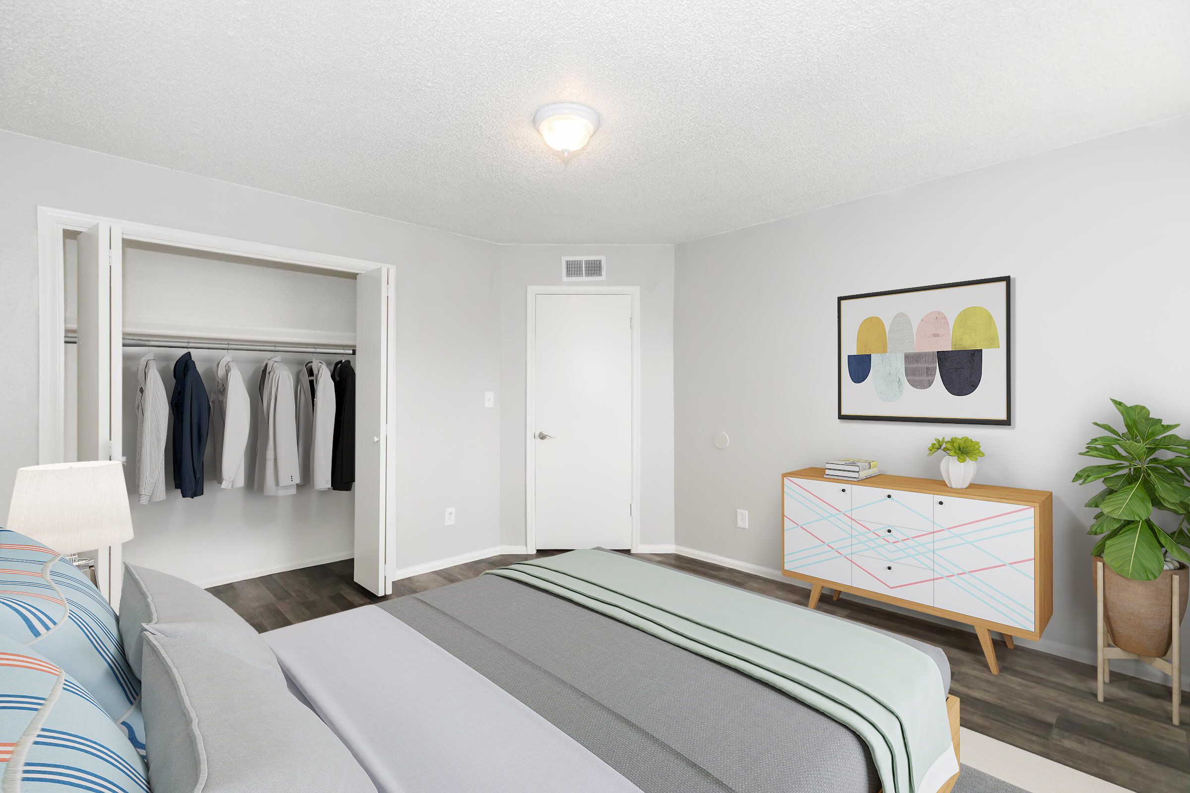 A modern bedroom featuring a neatly made bed with gray and striped linens, a stylish dresser with colorful accents, a potted plant, and a closet with neatly hung clothing. The walls are painted light gray, and there's an abstract art piece above the dresser. A light fixture adds warm illumination to the space.