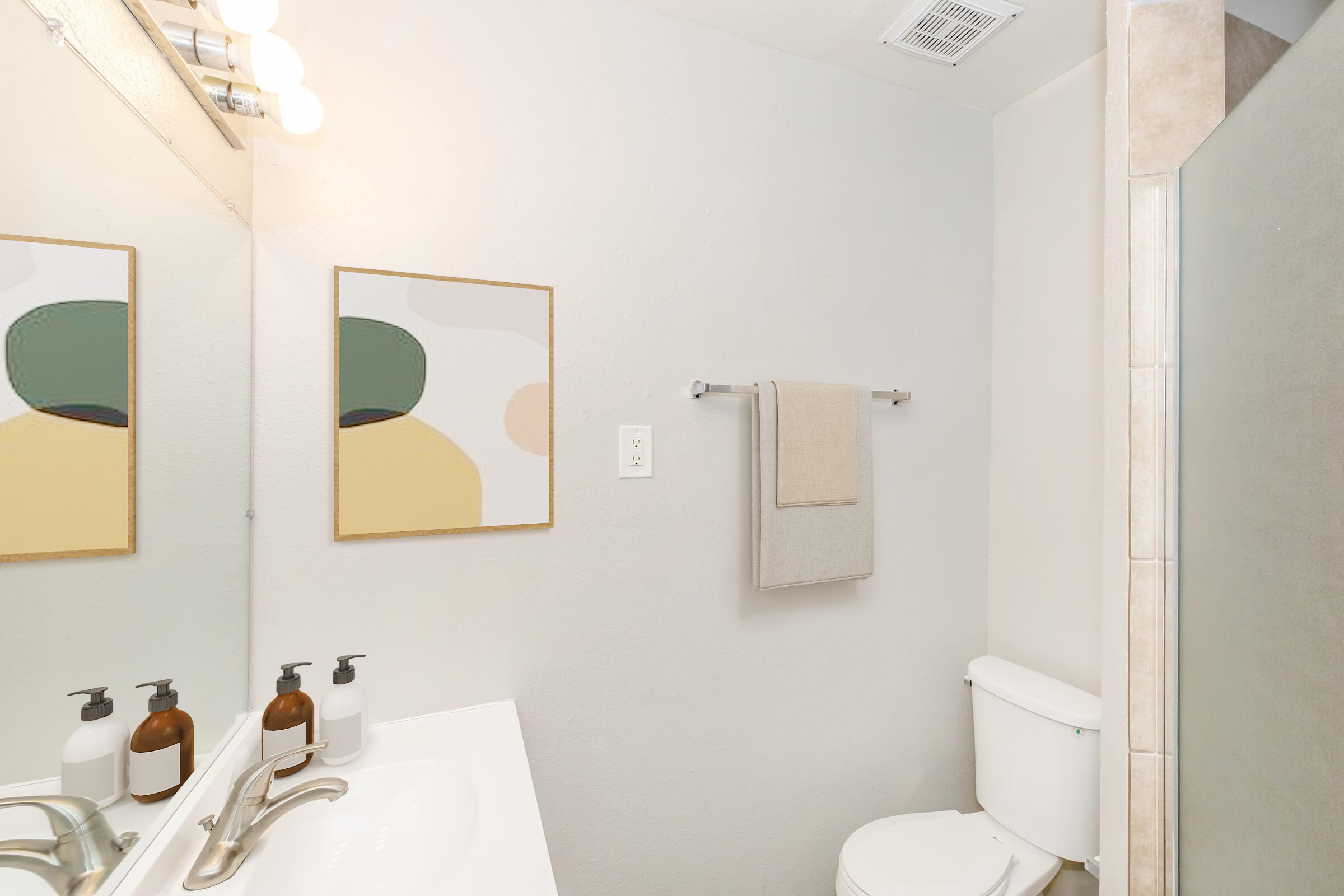 A clean and modern bathroom featuring a white sink with brown and white soap dispensers, a large mirror reflecting light, a beige towel hanging on a silver rack, and a toilet. Abstract artwork in muted colors adorns the wall, and the walls are painted a soft gray.