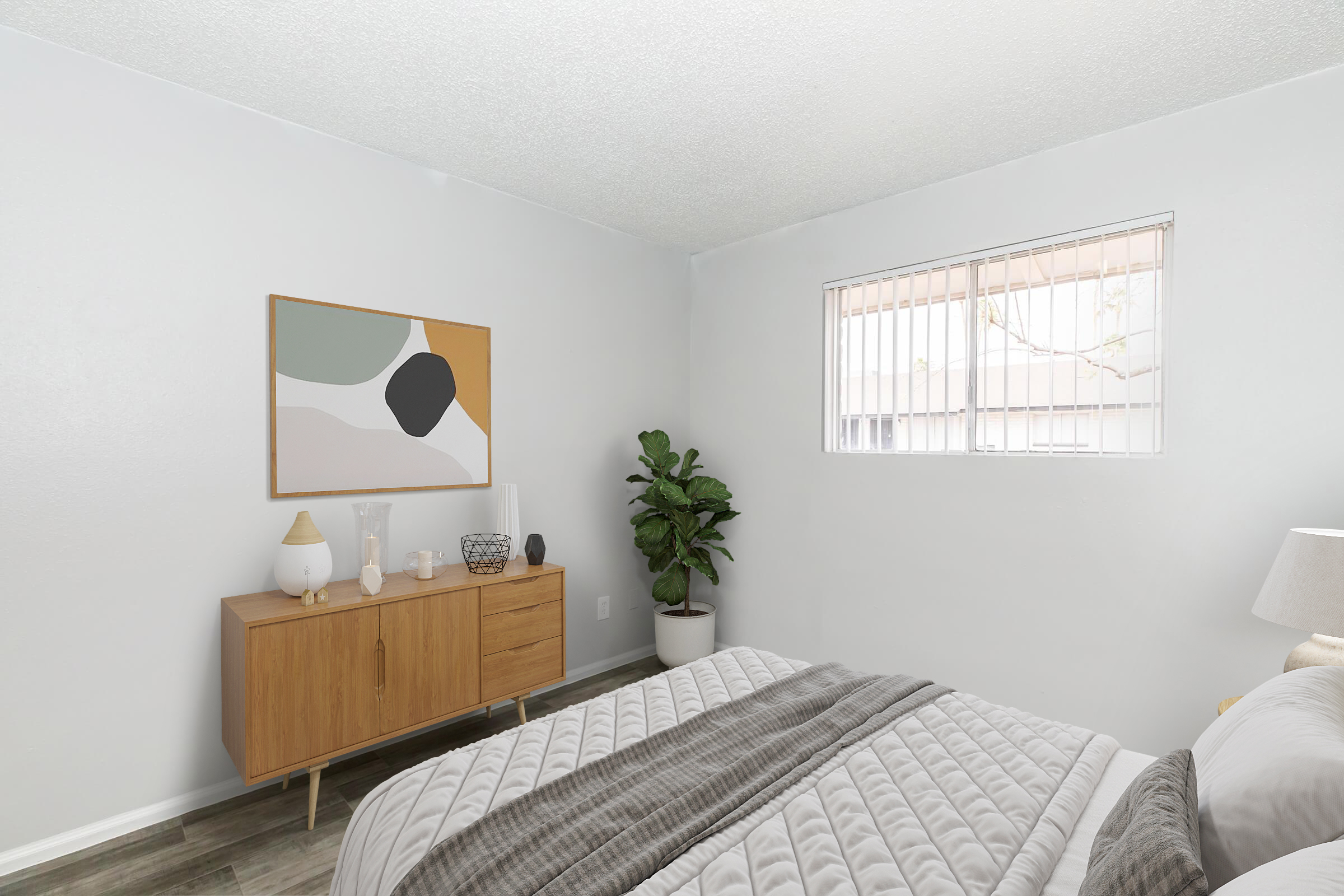 A cozy bedroom featuring a neatly made bed with gray and white bedding, a wooden dresser with decorative items, a large abstract wall art piece, and a potted plant by the window. Natural light filters through a simple window with horizontal blinds.