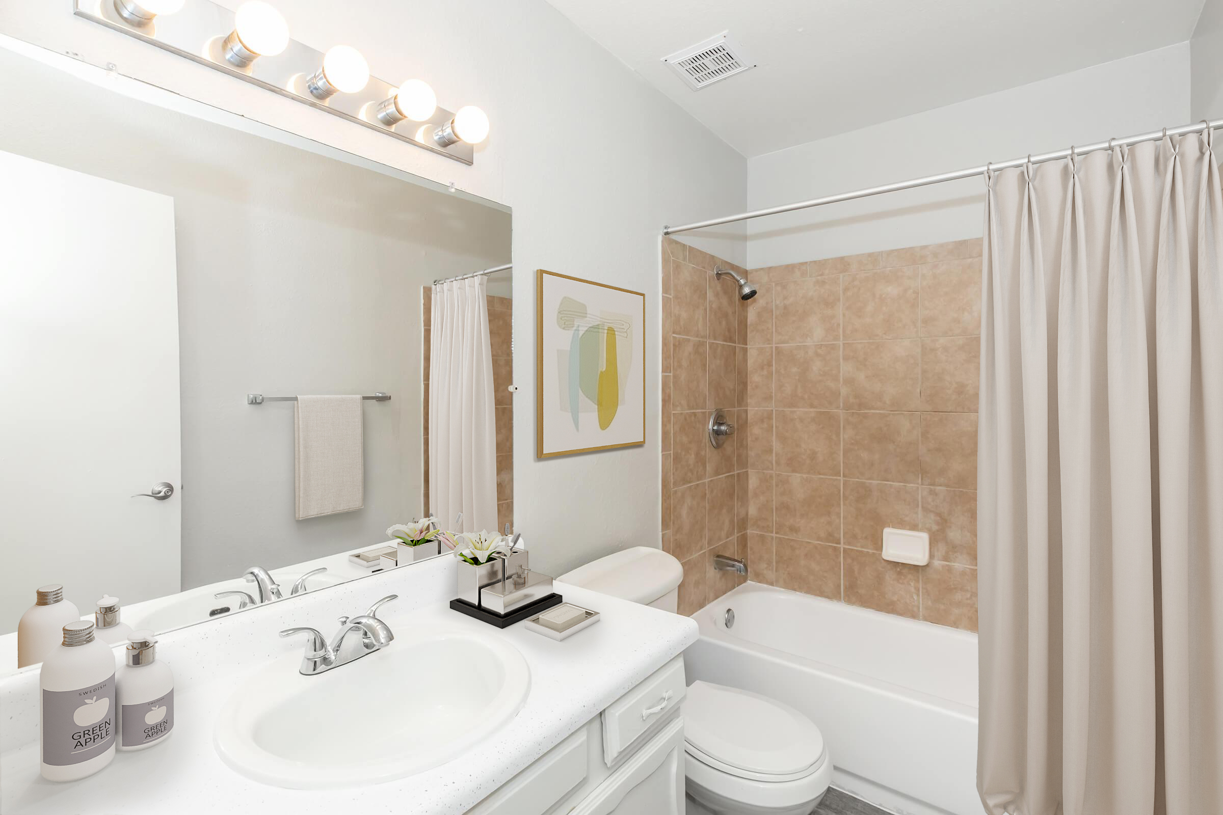 A clean, modern bathroom featuring a white vanity with a sink, two soap dispensers, and a decorative plant. The mirror above the sink reflects soft light from an overhead three-bulb fixture. There is a beige tiled shower with a curtain, an enclosed bathtub, and a woven towel hanging on the wall. A framed artwork adds a touch of color.