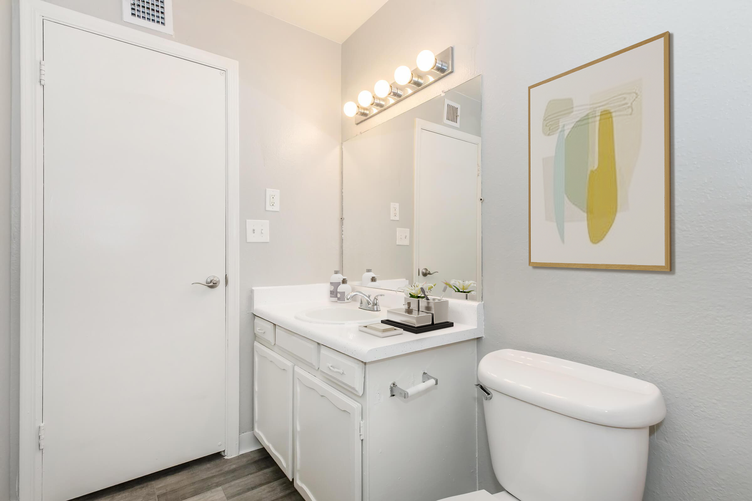 A modern bathroom featuring a white vanity with a sink, a wall-mounted mirror above, and bright lighting. A toilet is positioned next to the vanity. A small framed artwork is hung on the wall, and there are decorative items and plants on the countertop. The walls are painted in a light gray color.