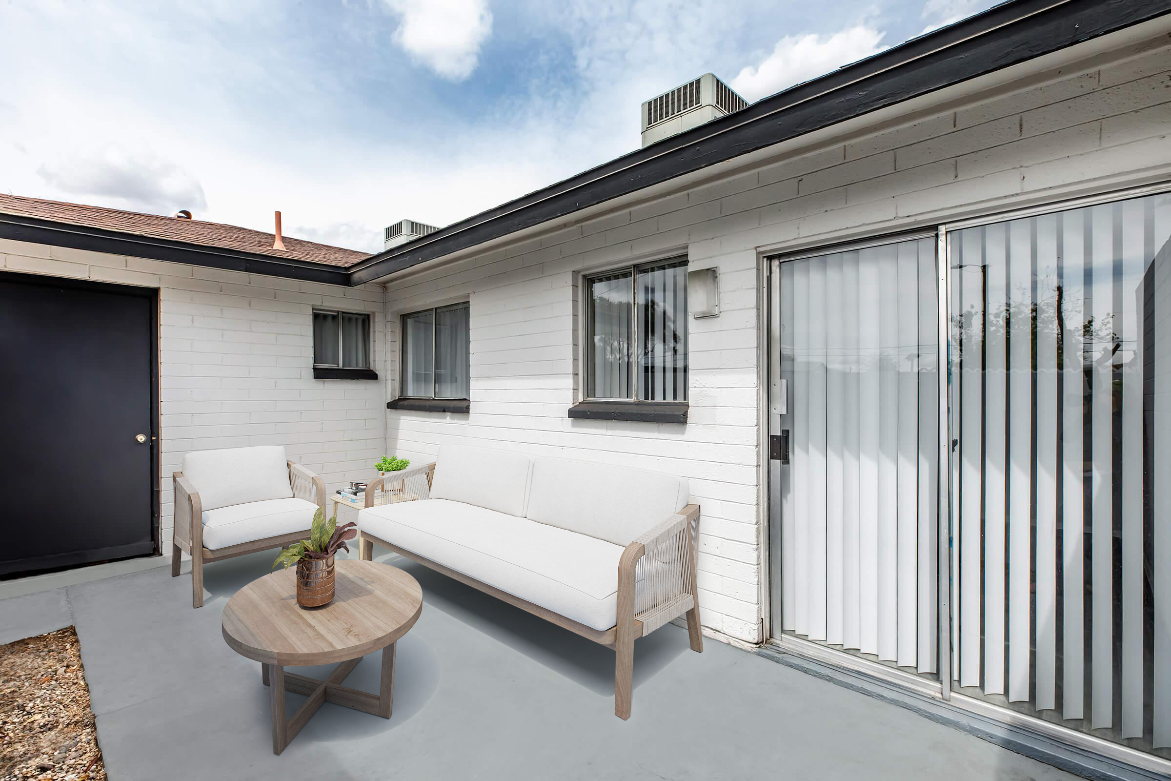 A modern outdoor patio area featuring a beige couch with matching chairs and a round wooden table. The space is surrounded by a white brick wall and has large glass sliding doors leading to the interior. The sky is partly cloudy, and there are potted plants for decoration.