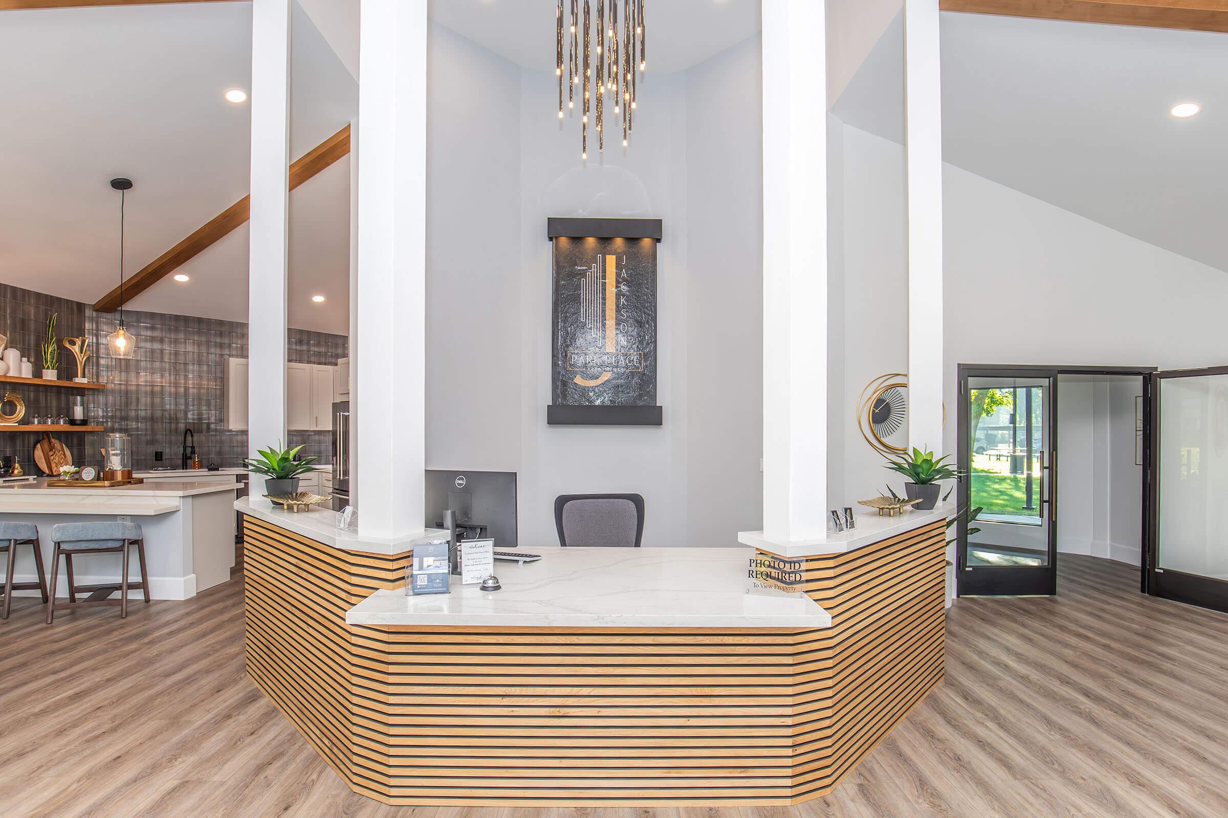 Modern reception area with a curved wooden counter, decorative wall feature, and stylish lighting. The space includes a seating area with bar stools and a kitchen-style counter in the background, featuring shelves and decor. Large windows allow natural light, creating a welcoming atmosphere.