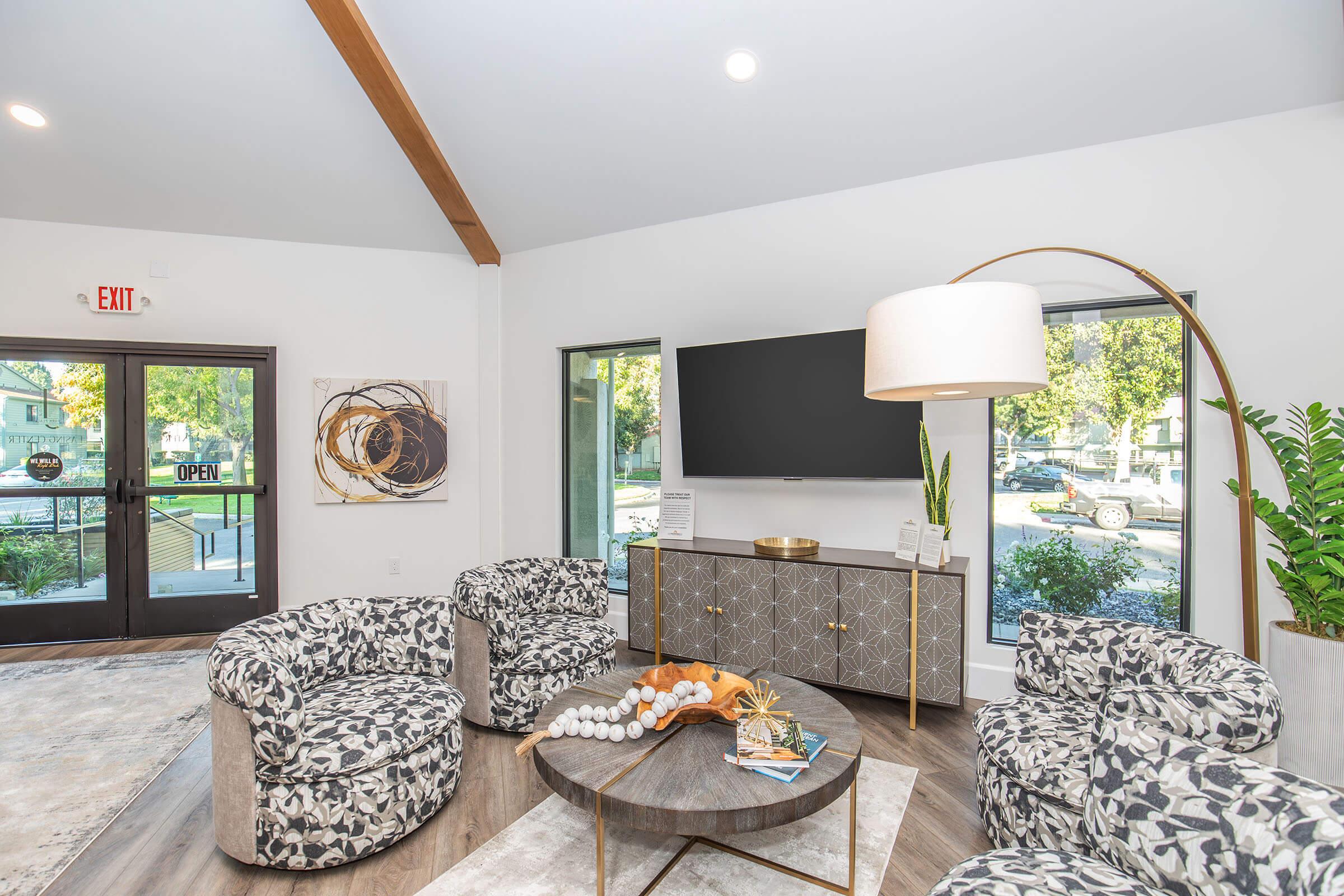 A modern interior living space featuring two large windows letting in natural light, a stylish coffee table with decorative items, and four patterned armchairs arranged around it. A flat-screen TV is mounted on the wall, and there is a large lamp illuminating the area. The entrance shows an "OPEN" sign.