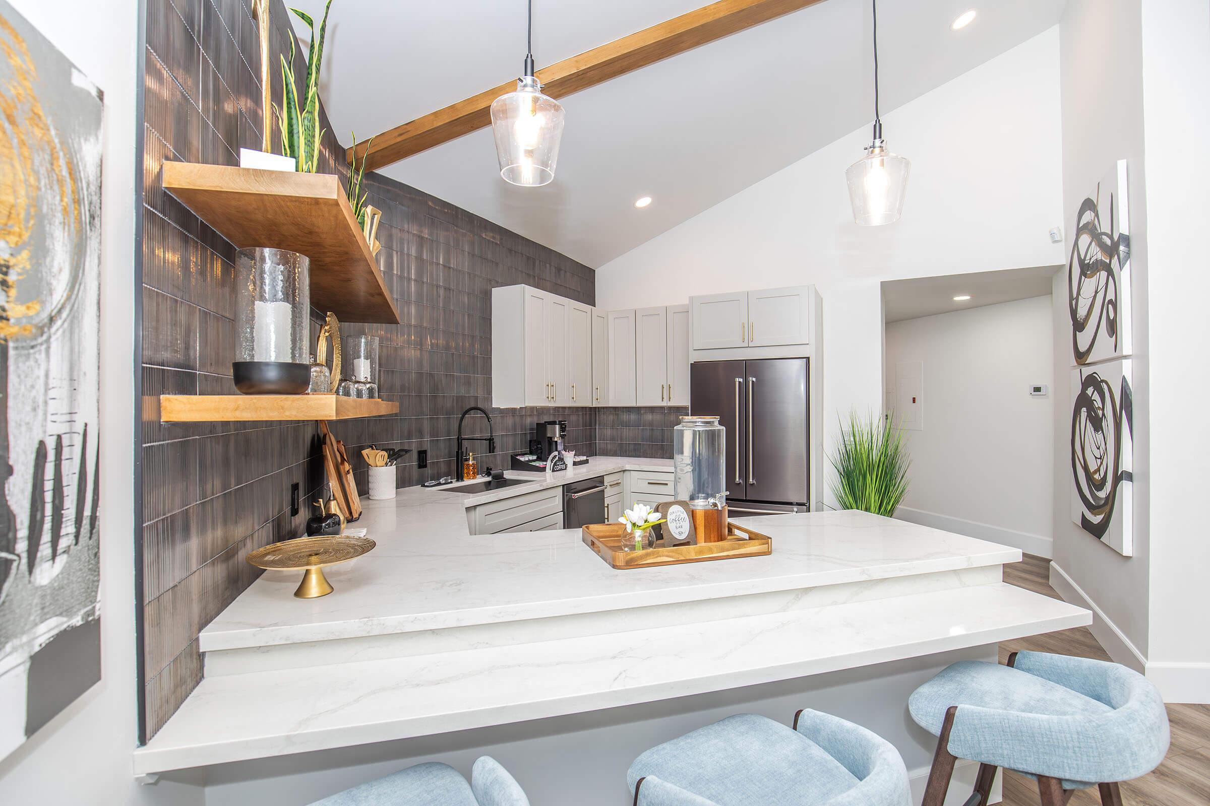 Modern kitchen featuring a white marble countertop with bar seating, wooden shelves displaying decor, and stainless steel appliances. The walls are adorned with gray tiles, and there is a touch of greenery. Pendant lights hang above the counter, creating a warm ambiance.
