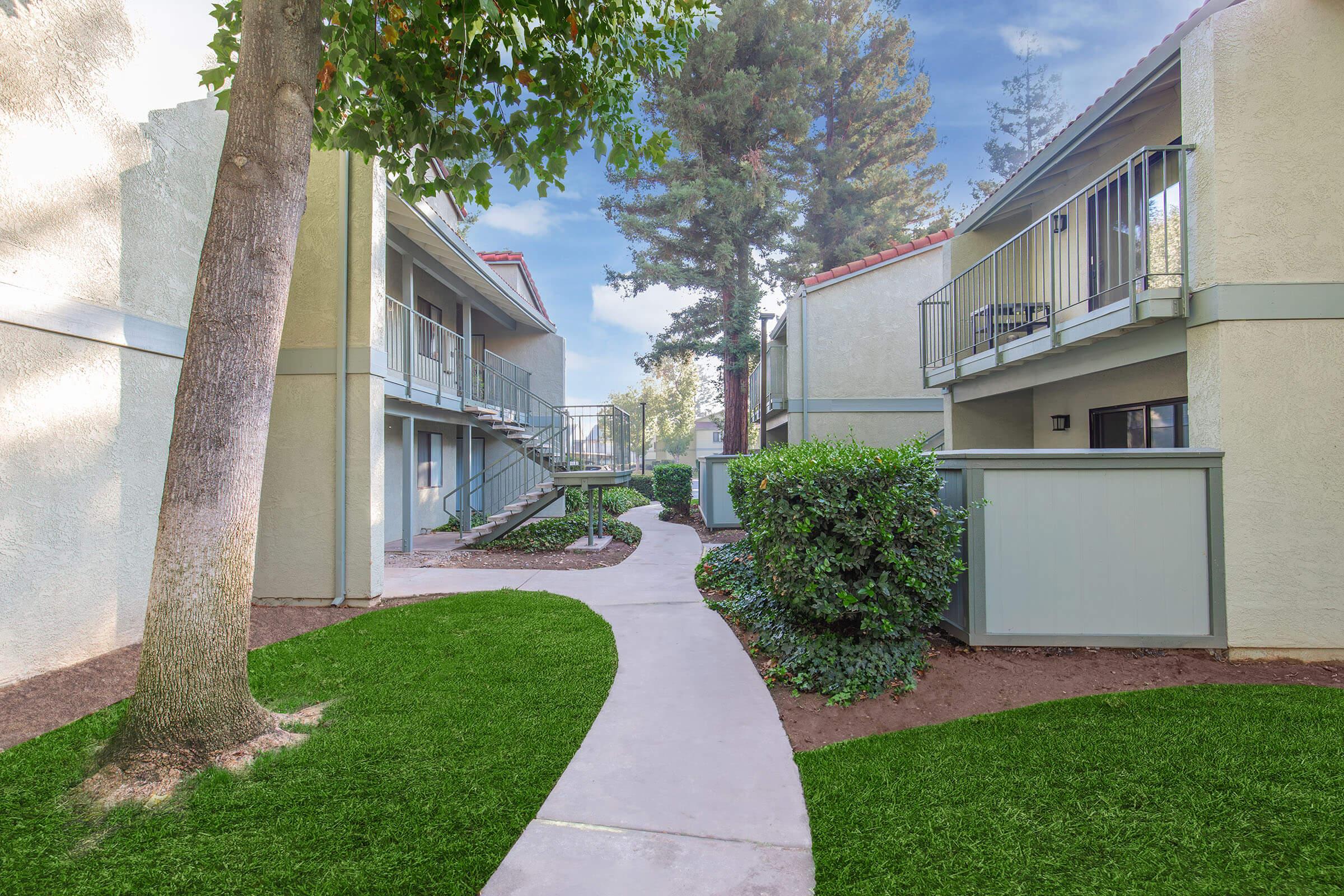 A landscaped pathway winds through a residential complex, flanked by two-story buildings. Lush green grass and trees provide a serene atmosphere, with balconies visible on the upper levels. The scene conveys a peaceful, well-maintained living environment.