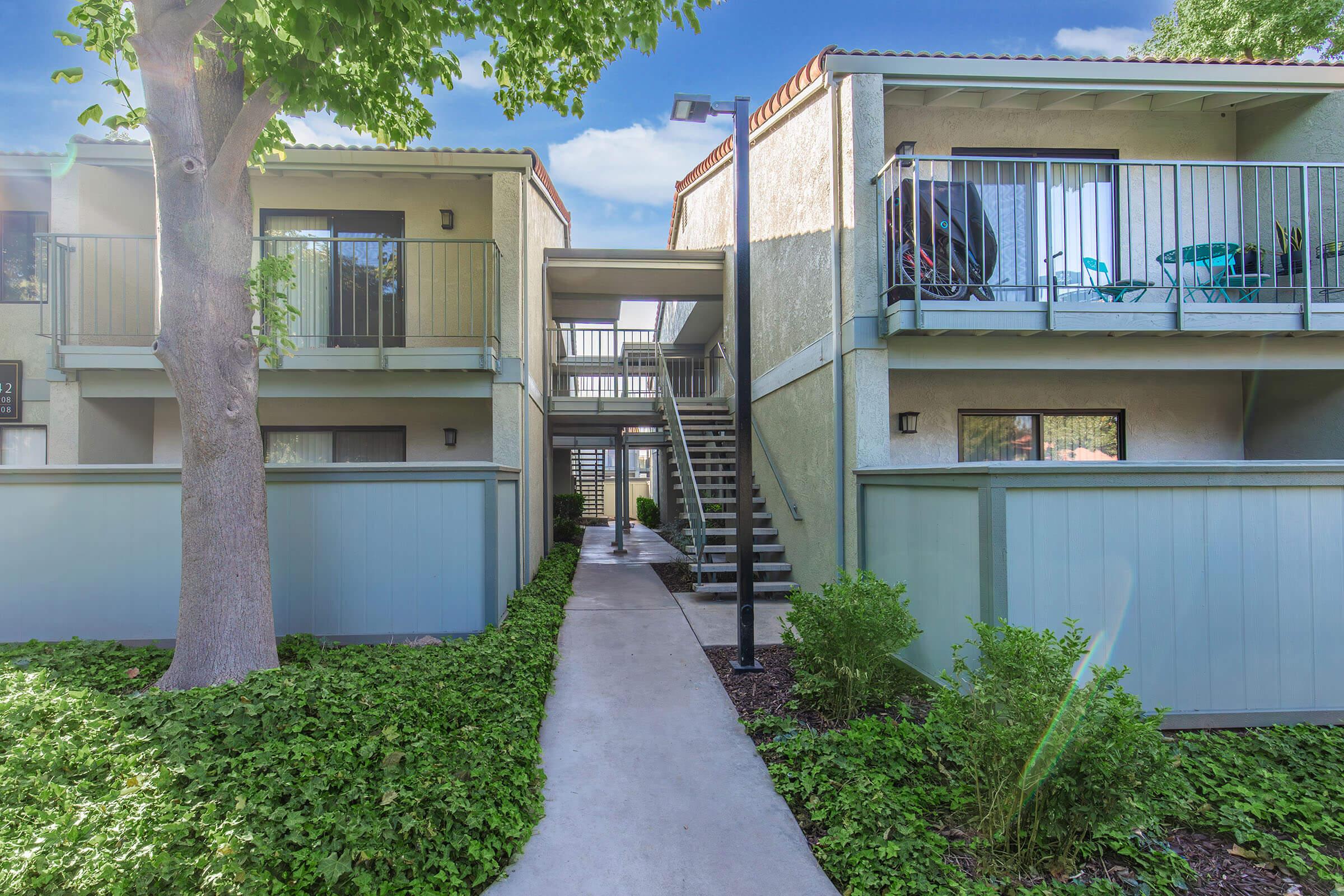 A view of a multifamily residential complex featuring two-story buildings with balconies, a tree on the left, and a well-maintained walkway lined with greenery. The path leads toward a staircase, with outdoor seating visible on the upper balconies. Bright blue sky in the background.