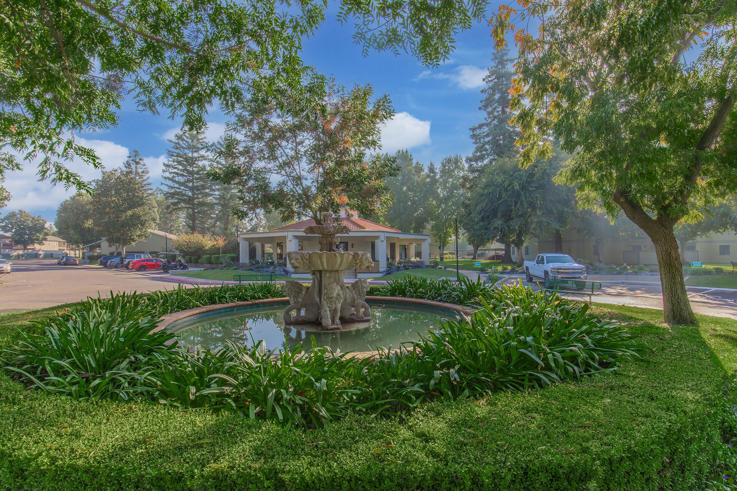 A tranquil scene featuring a decorative fountain surrounded by lush greenery and plants. The fountain is centered in a circular pond, with trees providing shade. In the background, a building with a sloping roof is visible, along with parked cars, creating a peaceful outdoor atmosphere.