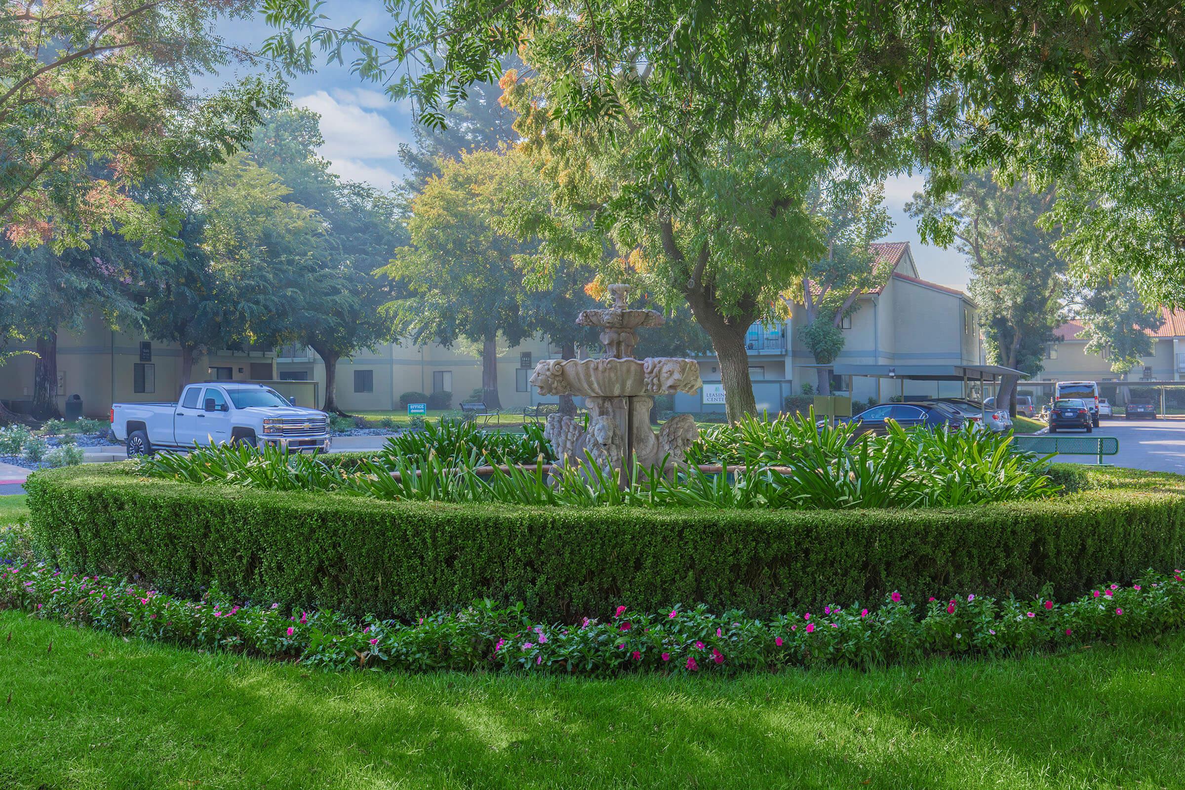 A lush landscaped garden featuring a stone fountain surrounded by neatly trimmed hedges and colorful flower beds. Tall trees provide shade in the background, with a glimpse of parked vehicles and buildings in a residential area. The scene conveys a serene and well-maintained outdoor space.