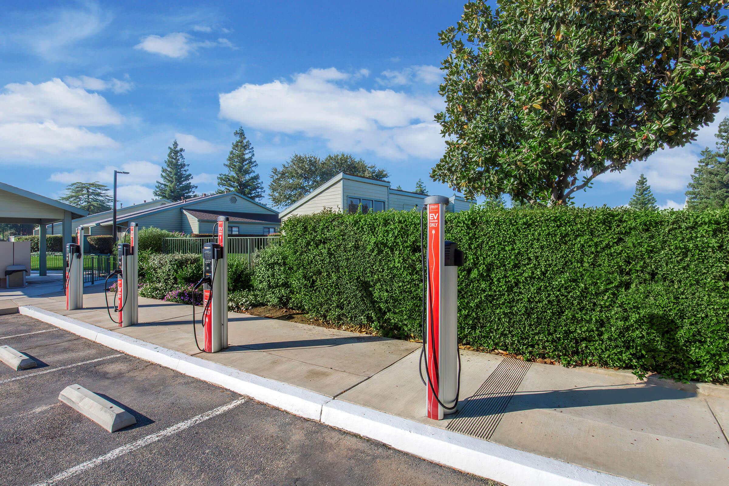Row of electric vehicle charging stations in a well-maintained parking lot, with a landscaped hedge and blue sky in the background. Nearby structures are visible, including a building and large trees, contributing to a clean and modern environment.