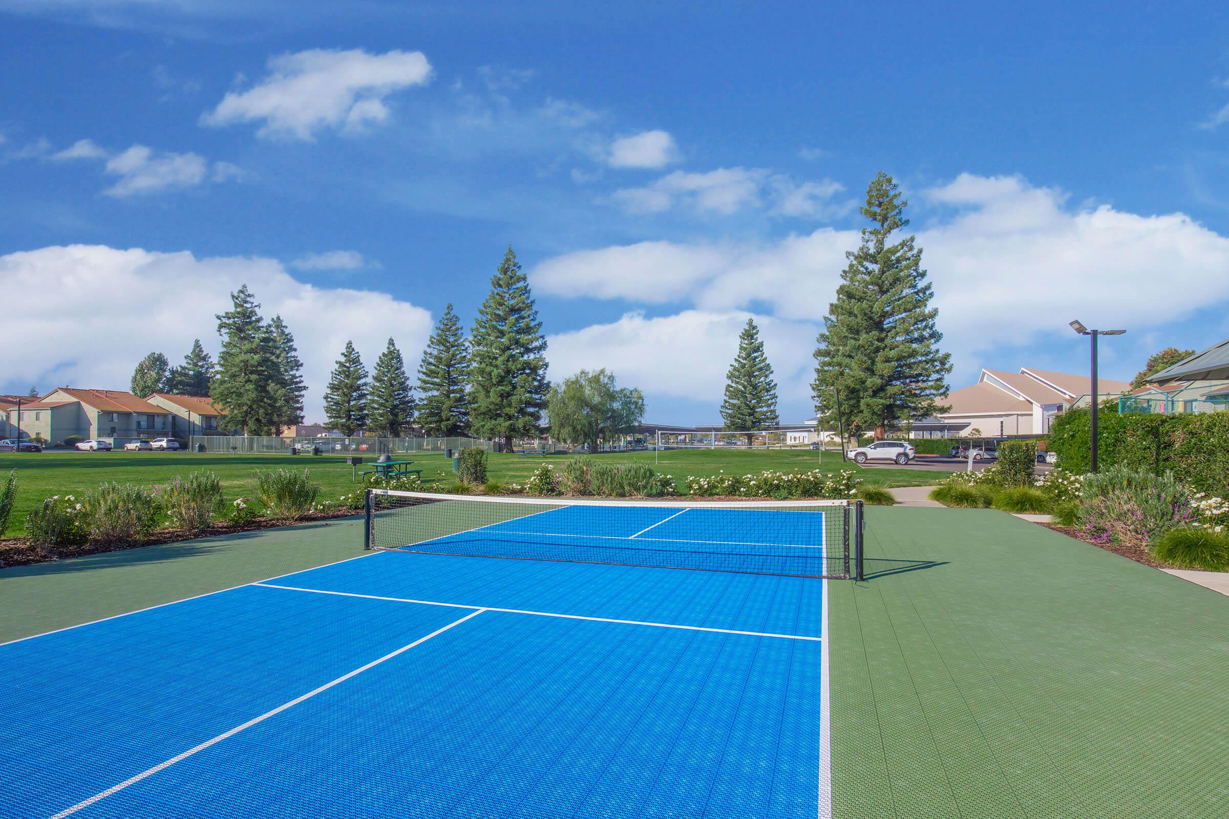 A bright blue tennis court with net in the foreground, surrounded by green grass and trees. In the background, there are residential buildings and a clear blue sky with scattered clouds. The scene is sunny, creating a vibrant and inviting atmosphere for outdoor activities.