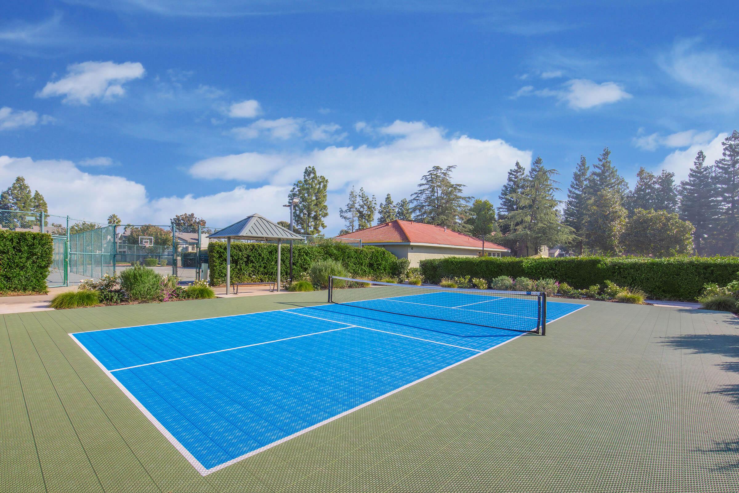 A bright outdoor tennis court with a blue playing surface, surrounded by lush greenery. In the background, there are two buildings, one a gazebo and the other a house, set against a clear blue sky. The court is ready for play with net in the center and neatly maintained surroundings.