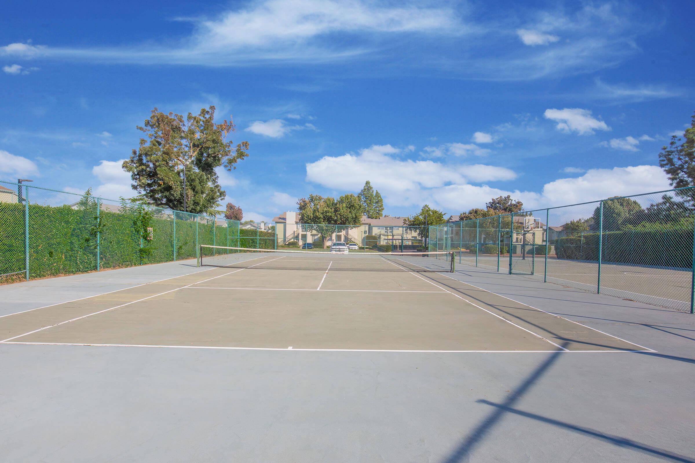 An empty tennis court surrounded by green fencing and trees, with buildings visible in the background under a blue sky with scattered clouds. The court has clear lines and is ready for play.