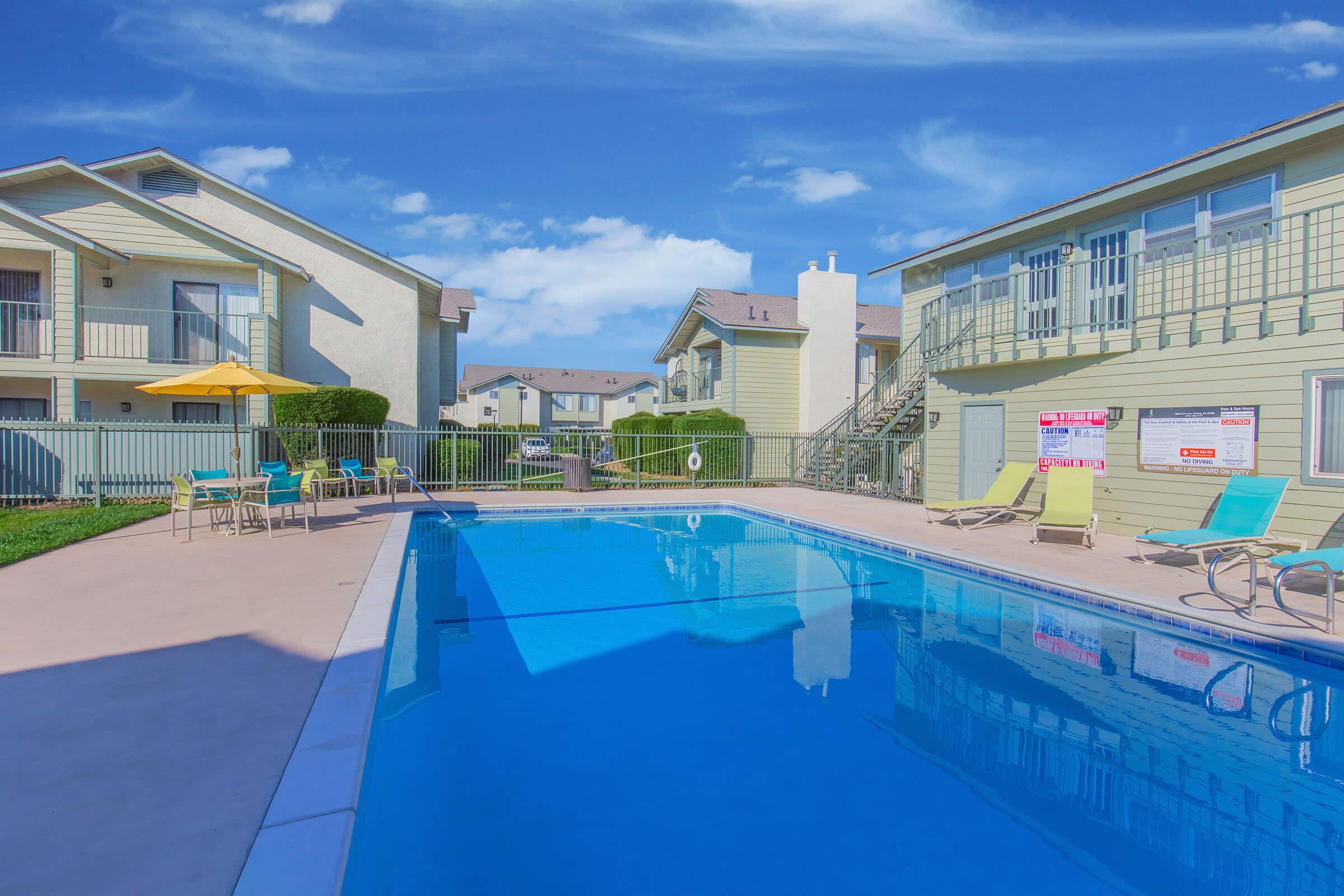 A view of a clear blue swimming pool surrounded by lounge chairs and tables, with a few umbrellas. The pool area is flanked by two residential buildings under a bright blue sky with scattered clouds. The setting appears inviting and well-maintained.