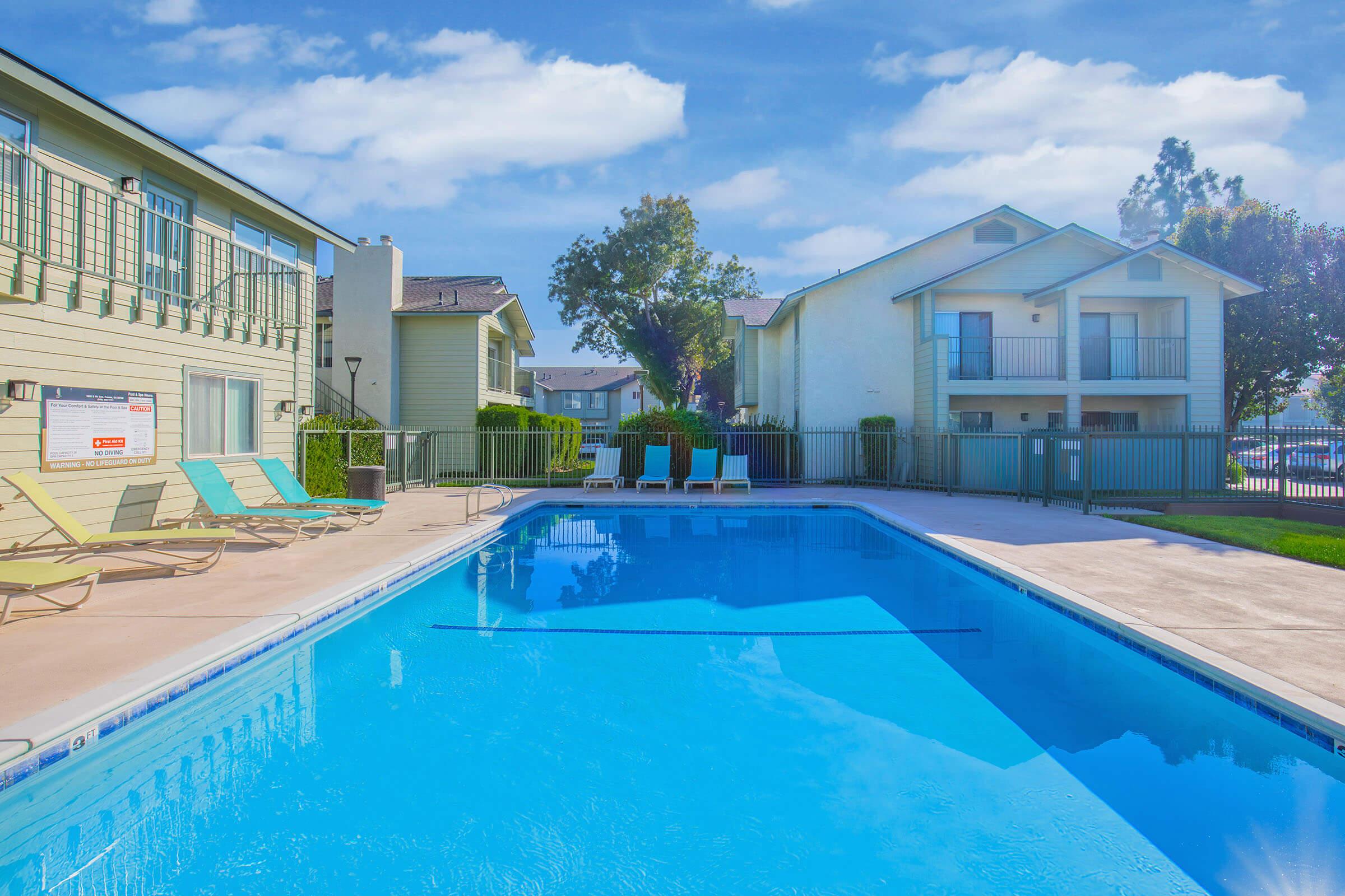 A clear blue swimming pool surrounded by lounge chairs, with two buildings in the background. One building has a fence and balconies, while the other is a single-story structure. Lush trees and a bright blue sky with a few clouds complete the tranquil scene.